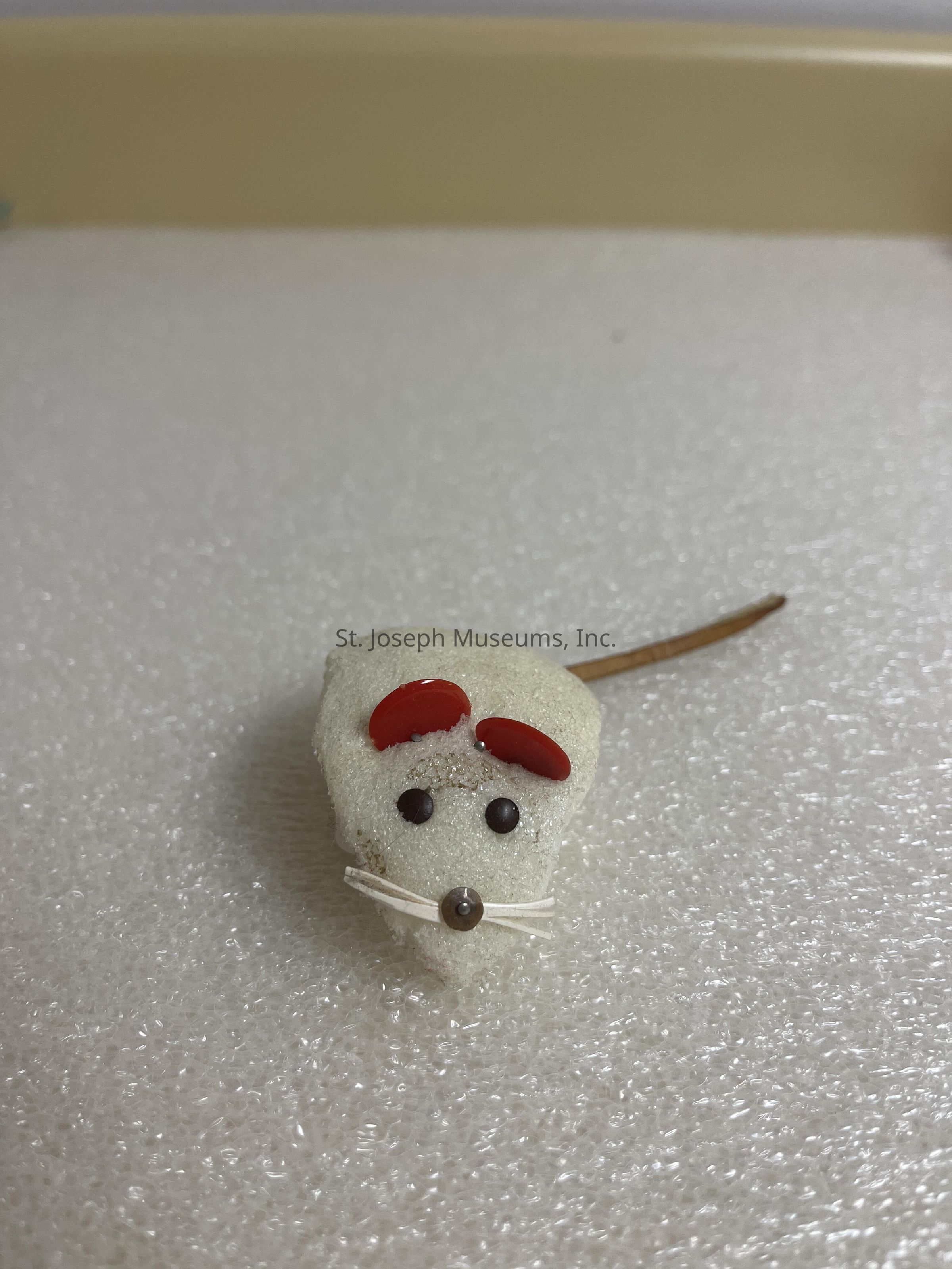The image shows a small, whimsical mouse crafted from a soft, sponge-like material, with red ears attached to the top. Its eyes are represented by two small, dark beads, and it has a tiny, rounded bead as a nose. Fine white strands protrude from either side of the bead, suggesting whiskers. The mouse's body rests on a slightly textured, white surface, and is completed by a long, thin tail extending from its back. The overall appearance is both simple and charming, capturing a playful, artistic interpretation of a mouse with a sense of handcrafted creativity.