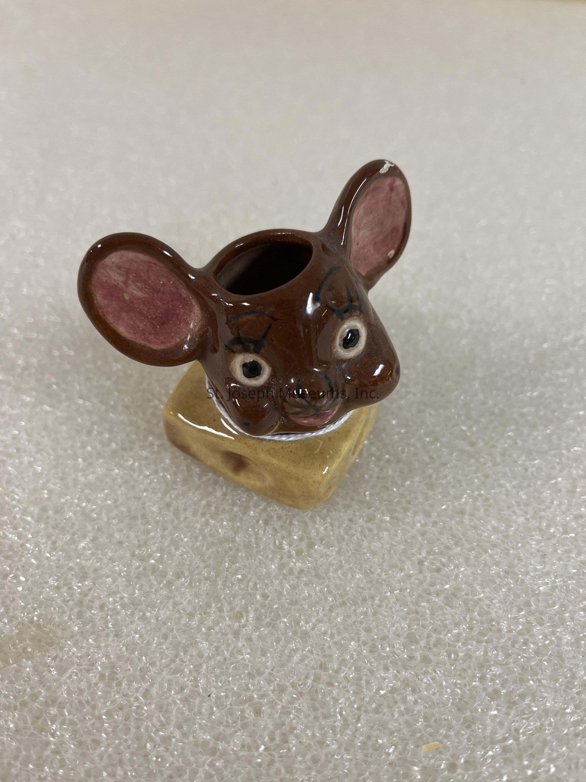 The image features a whimsical, ceramic figure designed to resemble a cute brown mouse with oversized ears, pink insides, and expressive wide eyes, set atop a small block that appears to mimic a piece of cheese with holes, all against a textured, light-colored background, possibly foam, which gives the figure an amusing and playful look, likely intended for decorative purposes in a kitchen or as a fun collector's piece, capturing a sense of humor and charm that would appeal to both children and adults who appreciate quirky or themed pottery items, characterized by its glossy finish and careful detailing that highlights its artistic craftsmanship.