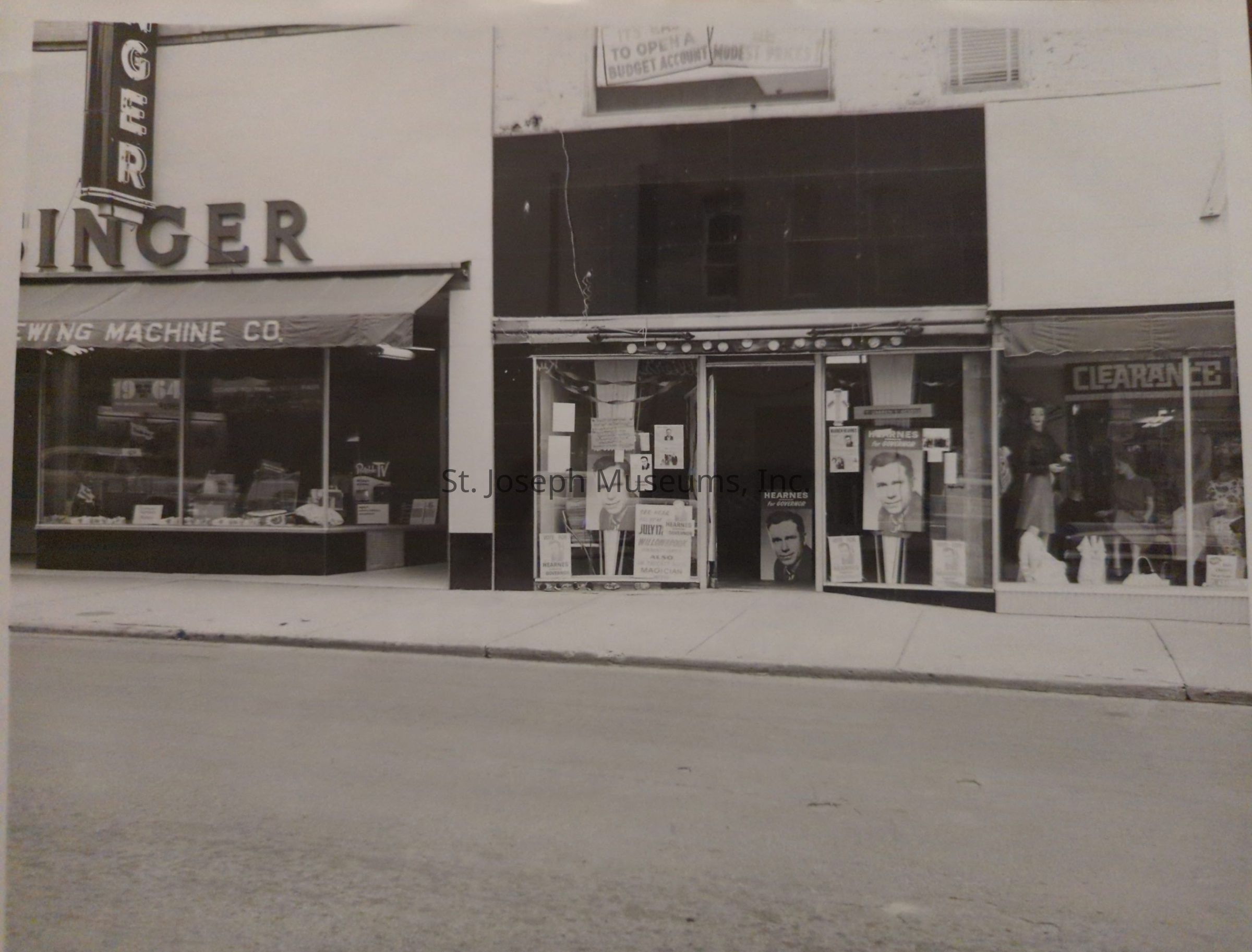Black-and-white photograph depicting a street scene of the Singer Sewing Machine Co. store with prominent signage and large display windows showcasing various products inside, while the adjacent store is adorned with numerous posters on the windows, one displaying a portrait labeled "Kearnes for Governor," with additional signage indicating a clearance sale, and a sidewalk stretches across the front, contributing to the vintage atmosphere captured in the photograph.