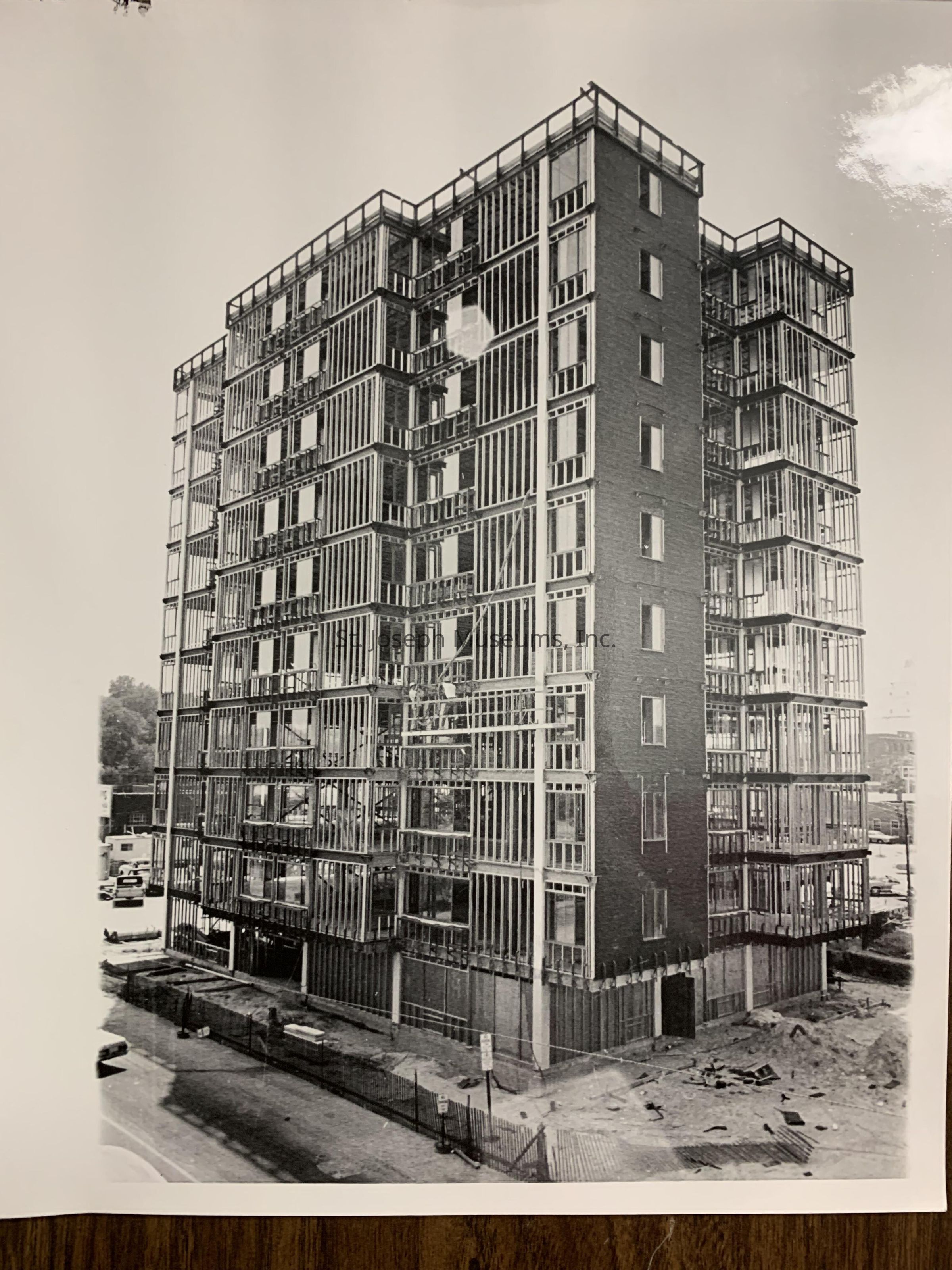 Black-and-white image depicting a St. Joseph Housing Project under construction, showcasing the VILLAGE EAST TOWERS APTS, at 1218 Village Drive, in an advanced stage of development. The structure's framework is visible, with steel beams and concrete forming the exterior. Scaffolding surrounds the building, indicating ongoing work, while construction materials and debris are scattered on the ground.