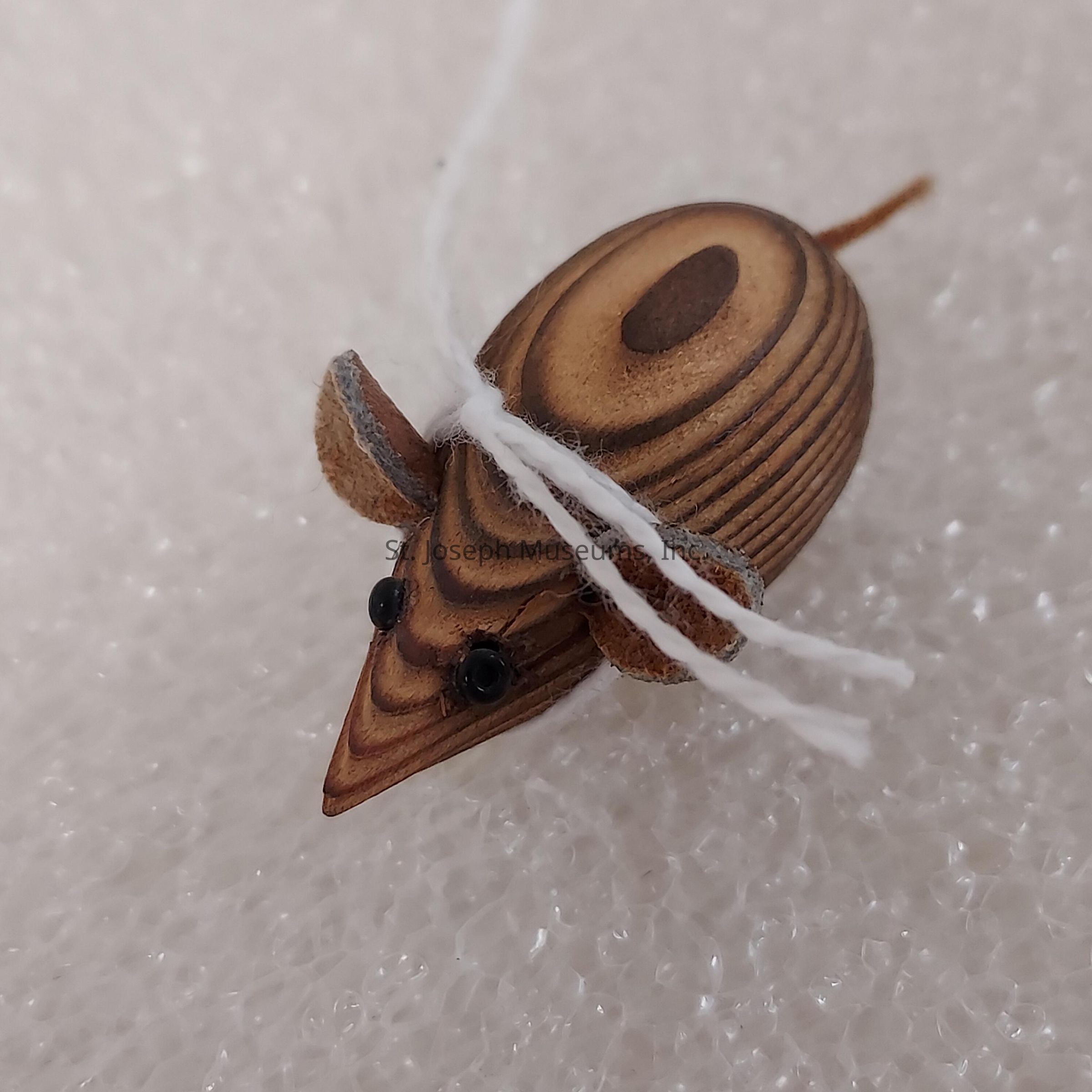 The image depicts a small, intricately carved wooden figurine in the shape of a mouse, featuring smoothly polished surfaces with natural wood grain patterns visible across its body, adding to its aesthetic charm. The mouse has two small, bead-like black eyes that give it an animated expression, while its pointed snout is characteristic of rodent designs in art and decorative crafts. Its tiny round ears are proportionate to its head, enhancing the lifelike quality of the figurine. A white thread loops around the mouse's body, adding a playful element to the design, lying on a textured background that contrasts subtly with the smoothness of the wooden piece. The mouse has a simplistic tail extending from its body, completing the miniature replica, and the craftsmanship demonstrates attention to detail and an appreciation for the simplicity of form, making it a delightful collectible or decor item.