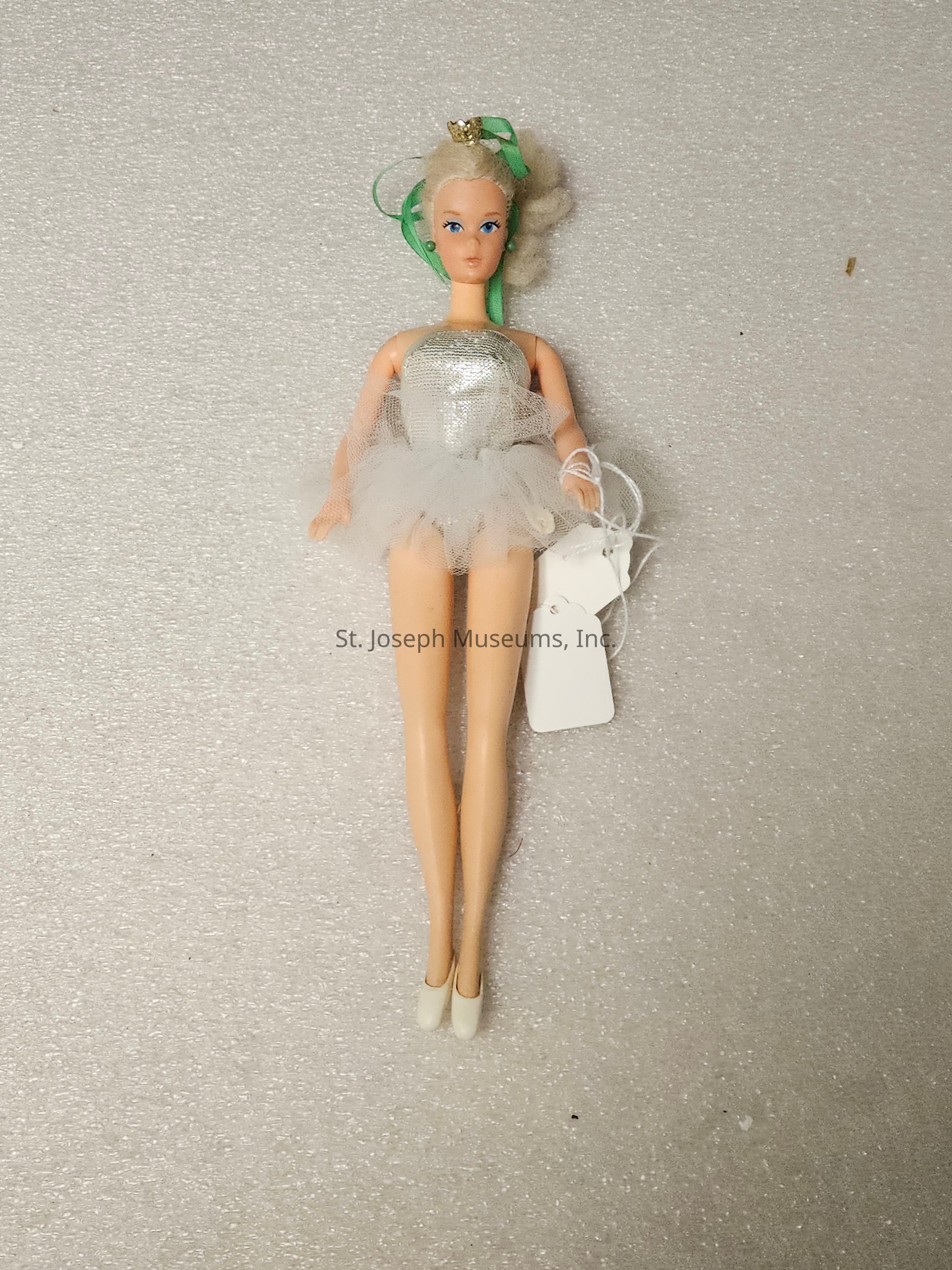 The image displays a doll with platinum blonde hair styled with a green ribbon and small crown. She wears a shiny silver bodice and a fluffy white tutu-style skirt, reminiscent of a ballet outfit. Her makeup includes bright blue eyeshadow, aligning with a classic doll aesthetic. The doll is positioned against a neutral, textured background. She is holding two white tags, attached with string, which might indicate a store or collector's item. Her feet are adorned with white ballet slippers, completing the ensemble. The overall appearance suggests elegance and a traditional ballerina theme.
