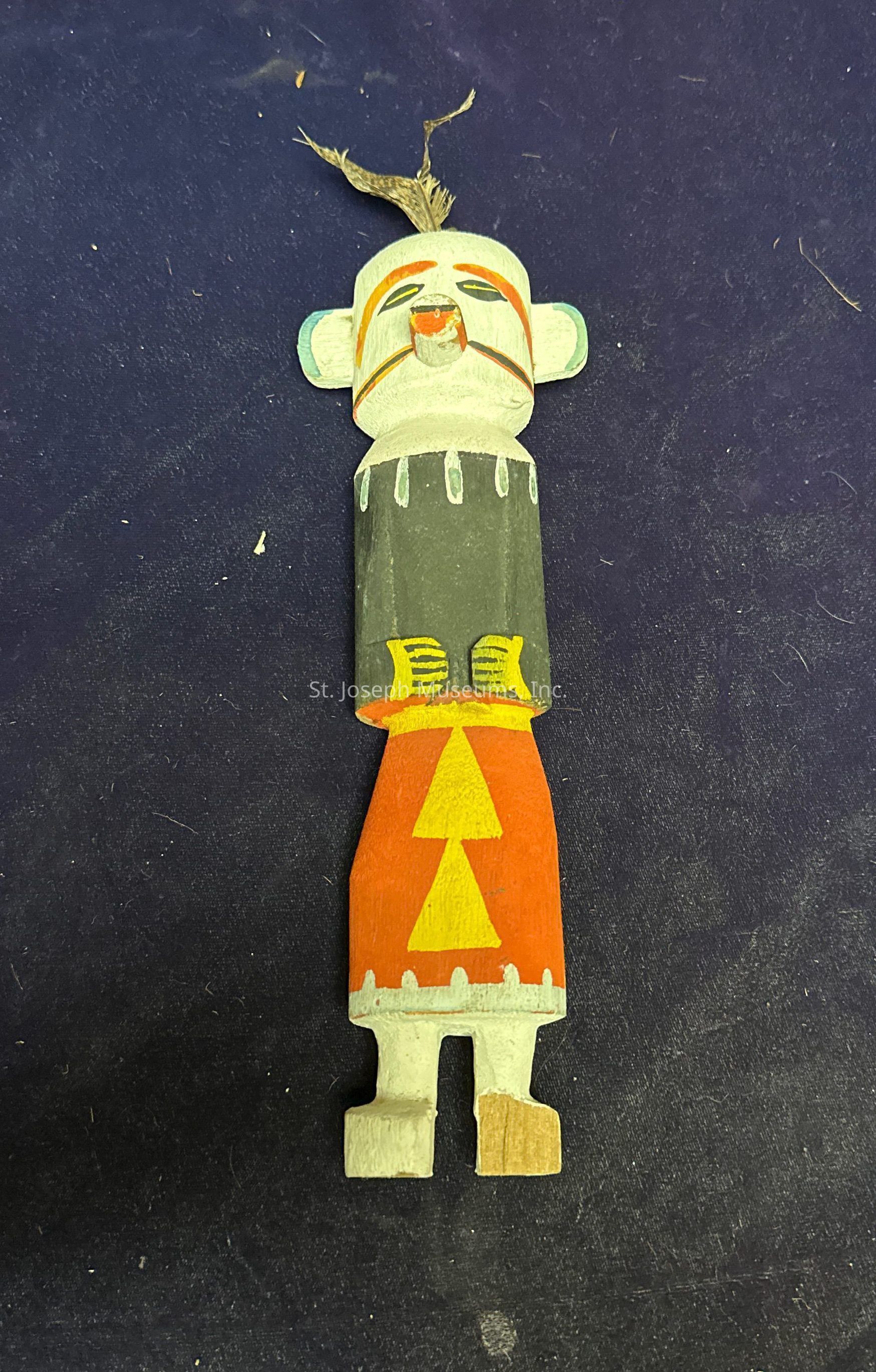 The image features a hand-carved wooden figure resembling a traditional kachina doll on a dark background. The figure is adorned with a combination of colors, including white, orange, yellow, and black, across its various body parts. Its head sports a stylized face with prominent eyes, a painted mouth, and a small tuft of natural-looking material on top, possibly representing hair or a decorative headpiece. The garment, painted in a combination of dark and vibrant hues, showcases geometric patterns and designs, including triangles and lines, adding a cultural and artistic touch to the figure. Overall, the craftsmanship highlights cultural symbolism and traditional artistry.