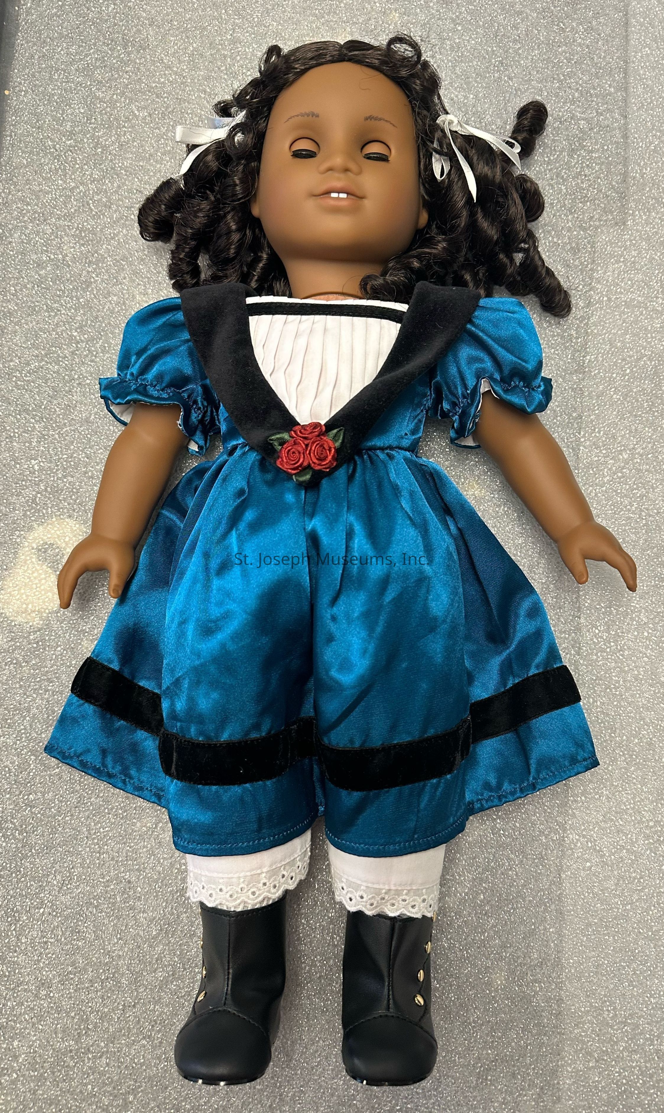 The image features a doll with curly hair adorned with white ribbons, dressed in an elegant blue gown with a distinctive black collar and puffed sleeves. The dress is accented with a red rose at the waist and trimmed with black velvet bands. It has a white pleated front, adding a touch of classical elegance. The doll is wearing black boots with decorative buttons, and under the gown, there are lace-trimmed white pantaloons visible. The overall attire and the doll's serene expression give it a vintage, nostalgic feel against the simple gray background.