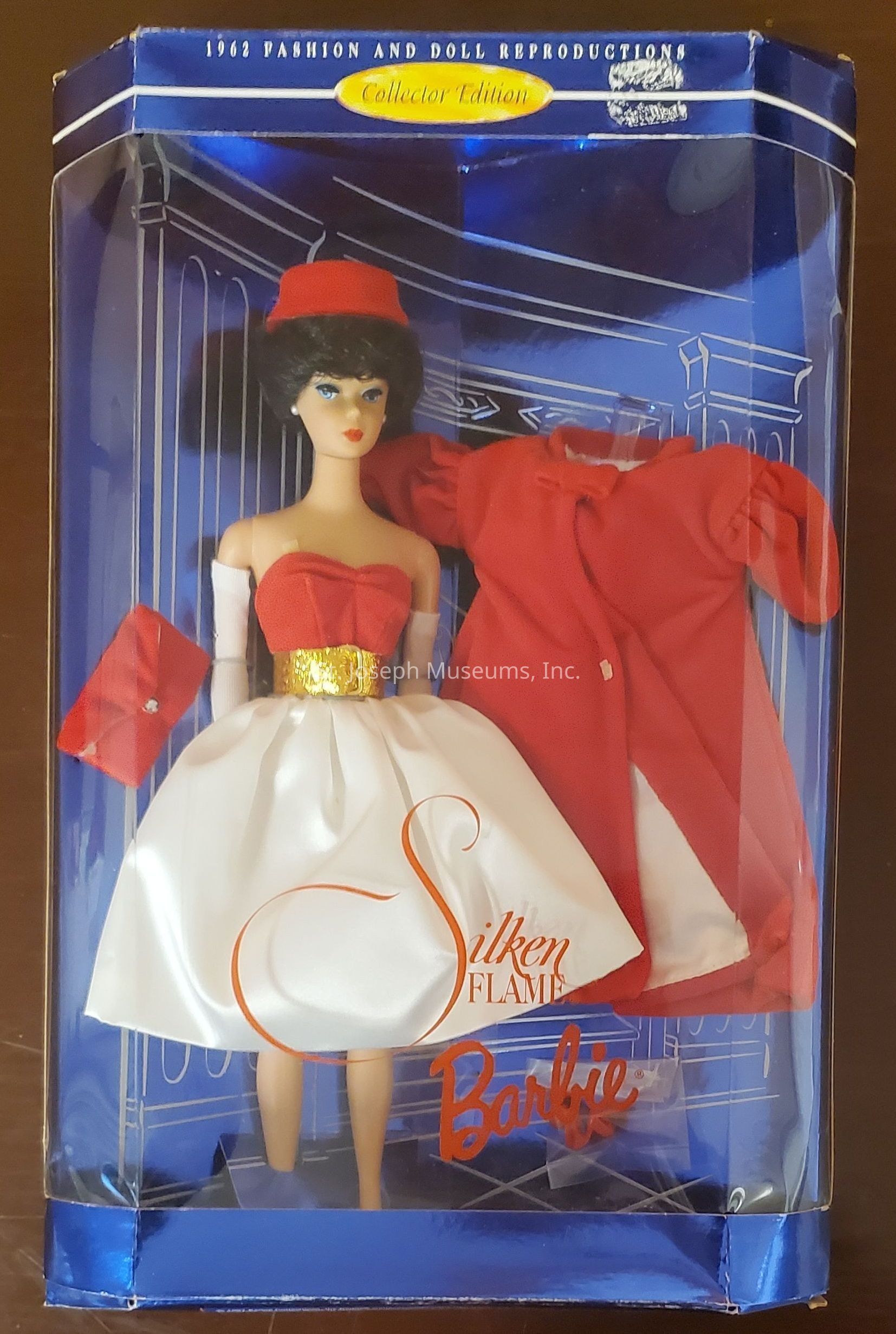 The image shows a collector edition Barbie doll in a display box marked "1962 Fashion and Doll Reproductions." The doll wears a red and white dress with a gold belt, accessorized with long white gloves, a red hat, and holds a red clutch. A red coat is also included in the packaging, complementing the elegant ensemble. The display box has a blue background with decorative line art, and the phrases "Silken Flame" and "Barbie" are prominently featured on the front of the clear packaging. This classic design highlights vintage fashion trends from the early 1960s.