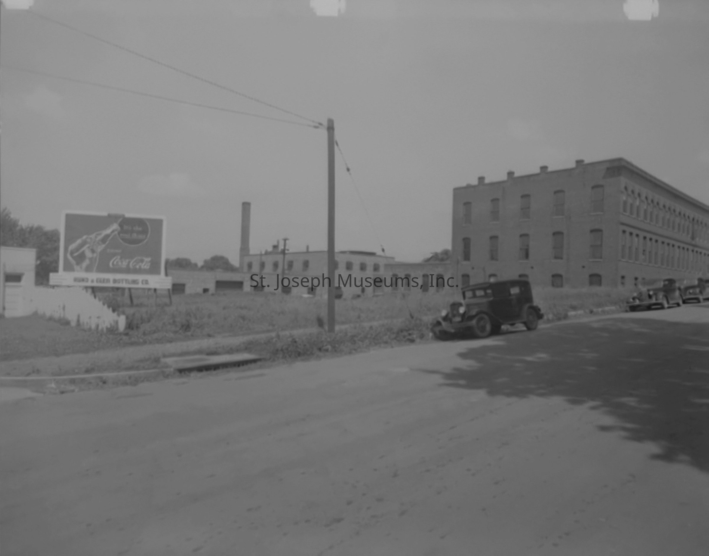 Black-and-white image depicting the exterior of the Sterling Casket Company located on 12th Street, south of Penn, in St. Joseph, Missouri. The large brick building dominates the right side of the scene, showcasing its multiple rows of windows. In front of the building, vintage automobiles are parked on the side of the road. To the left, there is a Coca-Cola advertisement featuring the slogan, "It's the real thing." The area surrounding the building is somewhat overgrown, with a utility pole and sewer drain visible.
