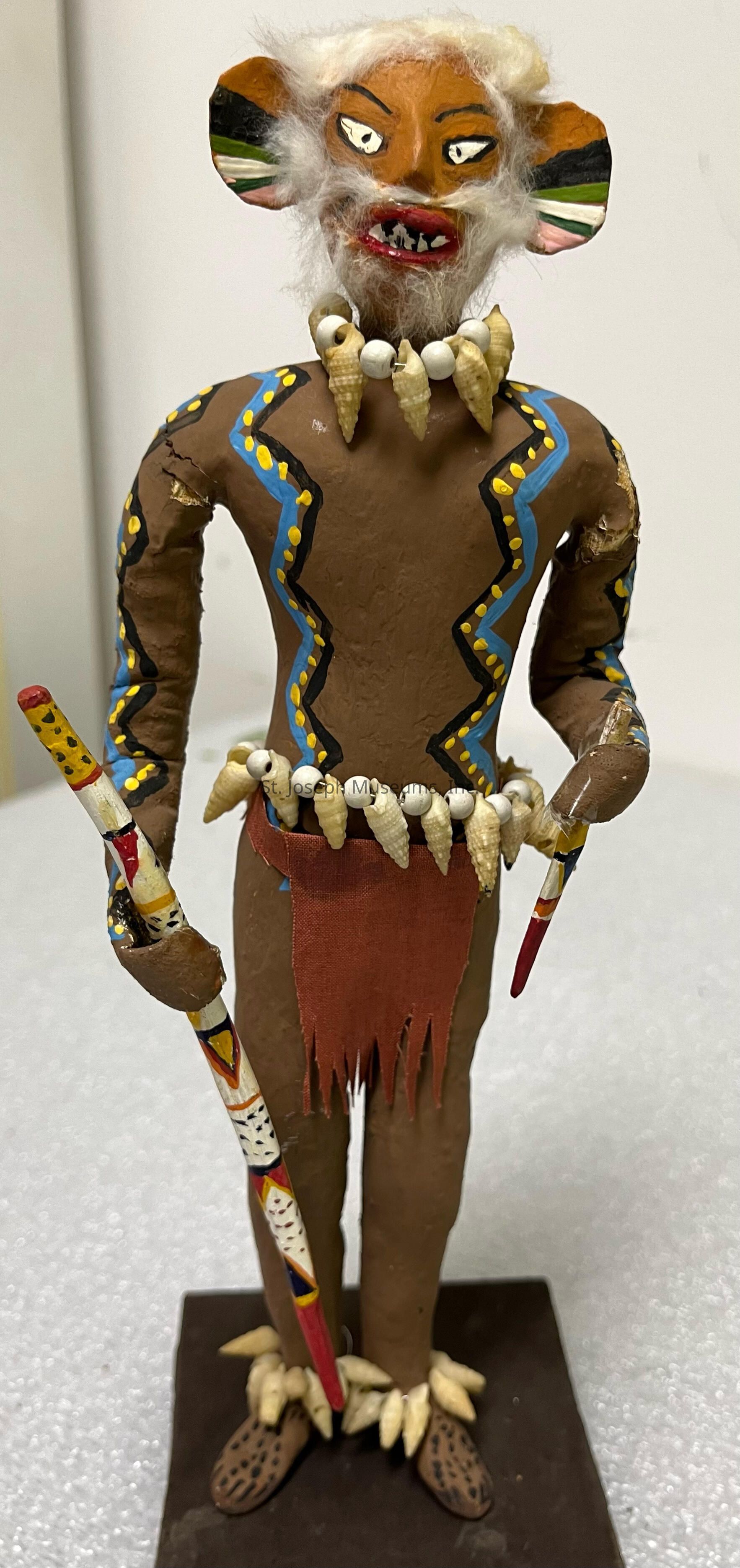 The image shows a handcrafted figurine with an intricate design featuring a humanoid figure adorned with a decorative, brown body suit decorated with blue and yellow patterns. The figure has a fierce expression, prominent fangs, and large ears, with white painted eyes and a detailed mouth area intricately painted to suggest sharp teeth. The figure holds a colorful staff in its right hand, painted with red, white, yellow, and black stripes and patterns. Around its neck, it wears a necklace made of small bead-like shapes, and its waist is adorned with a belt of shell-like objects along with a red, fringed cloth hanging in front. The legs and feet are also detailed, incorporating a pattern of spots on its shoes, and the entire figure stands on a rectangular base, suggesting a ceremonial or tribal theme, and is likely crafted from a material resembling clay or papier-mâché.