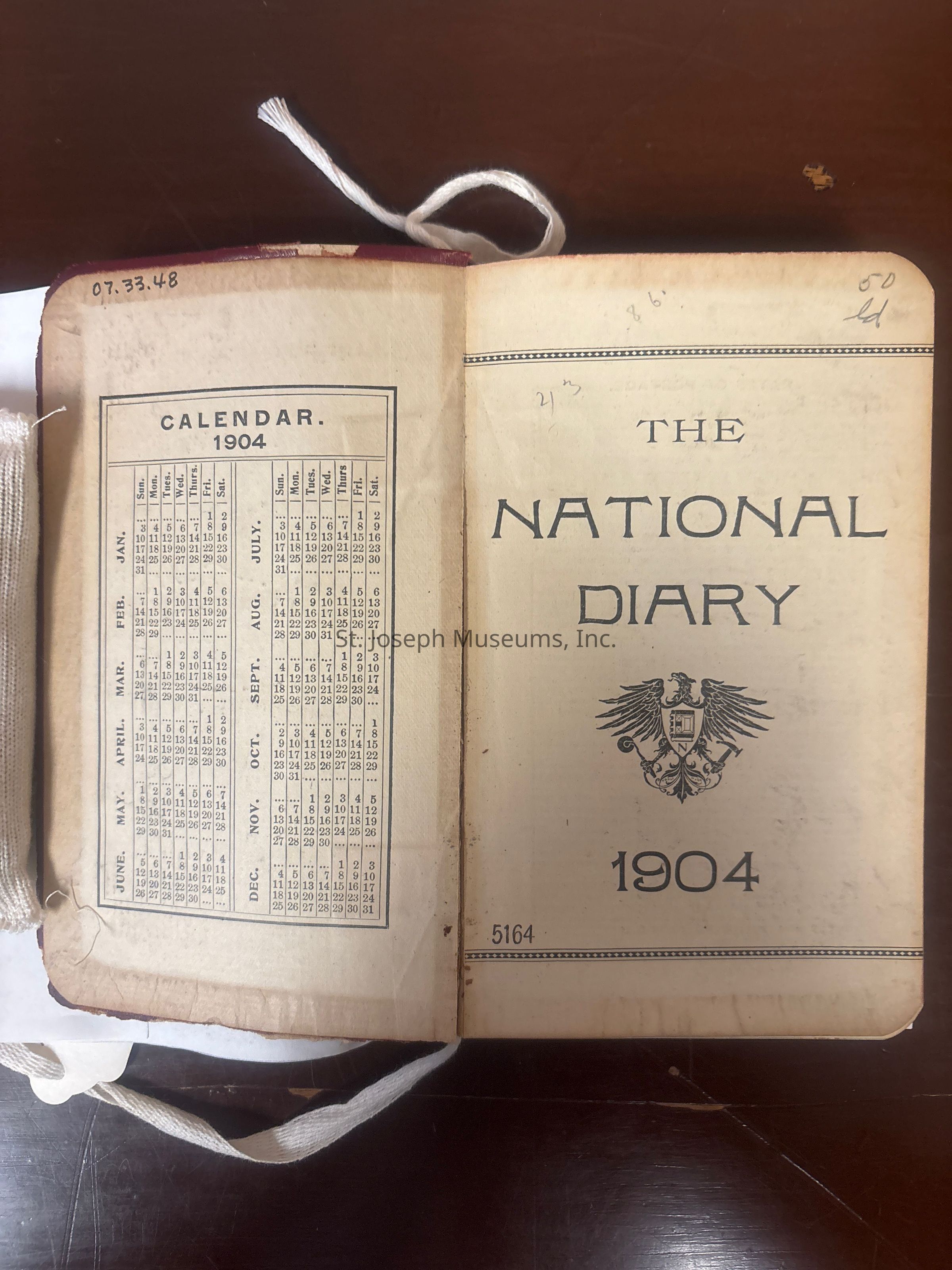 The image depicts an open diary titled "The National Diary 1904," displaying its interior pages. The left page features a printed calendar for the year 1904, organized month by month in a grid format, with handwritten numbers at the top corners. On the right page, the title "THE NATIONAL DIARY 1904" is prominently displayed, alongside an emblematic design featuring an eagle with a shield. The diary seems to have an aged and slightly worn appearance.