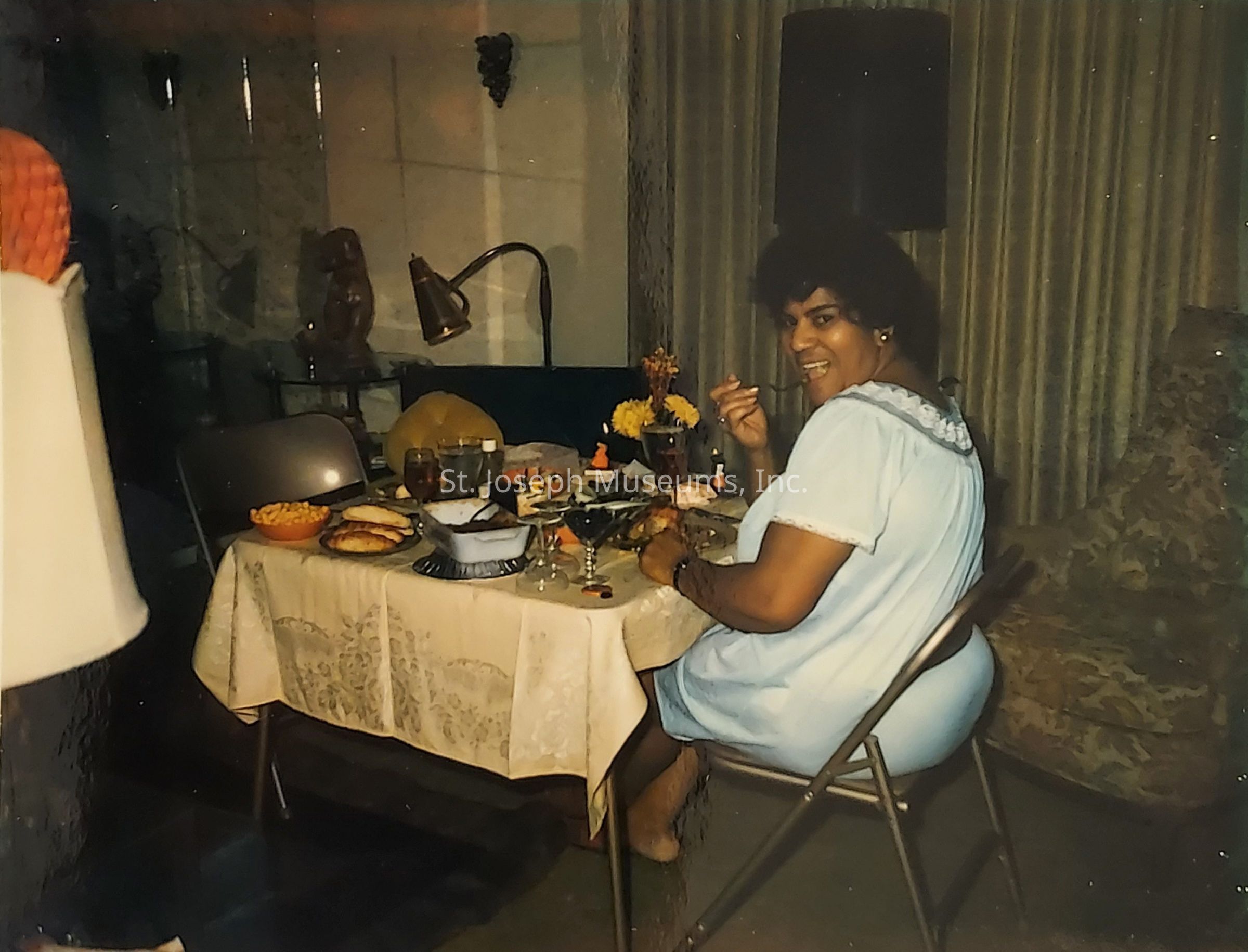 A woman wearing a light blue dress sits at a dining table, enjoying a meal and smiling at the camera. The table is covered with a lace tablecloth and is set with various dishes, including a bowl of snacks, drinks, and other food items. The room features subdued lighting, with curtains drawn in the background and a lamp providing a warm glow. A decorative figure and wall art add to the cozy ambiance of the setting. The woman appears relaxed and content, enhancing the welcoming atmosphere of the scene.