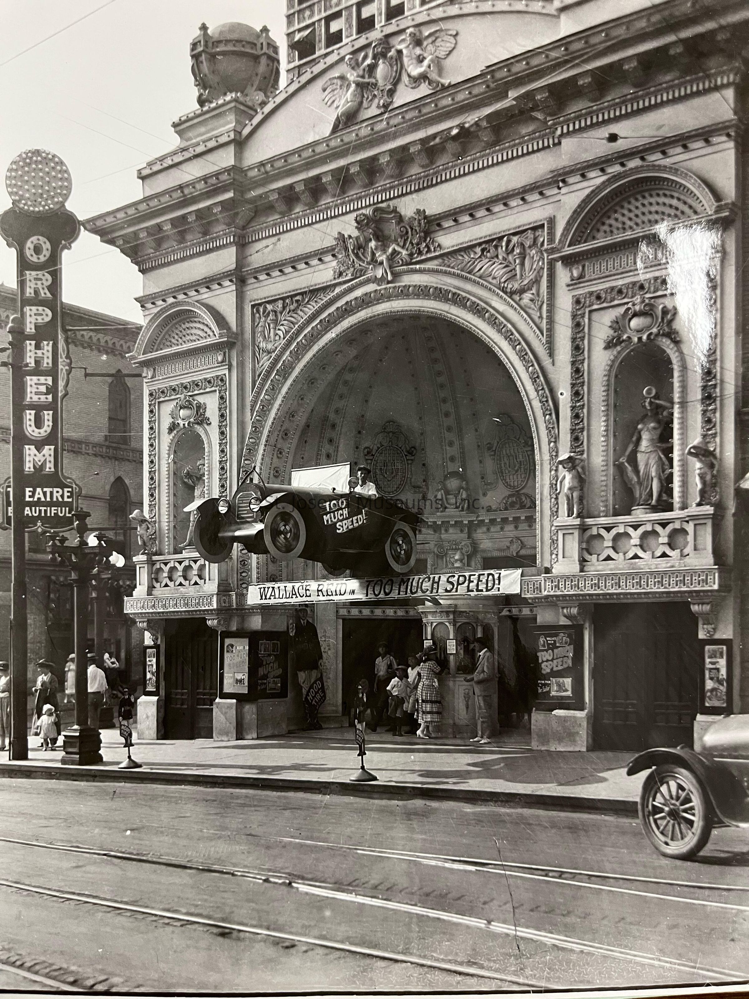 Black-and-white image depicting the ornate exterior of the Orpheum Theatre, with a large eye-catching sign and elaborate architectural details. A suspended vehicle above the entrance advertises "Too Much Speed," a film, drawing attention from pedestrians. People gather around the entrance, creating a lively street scene from 1925.