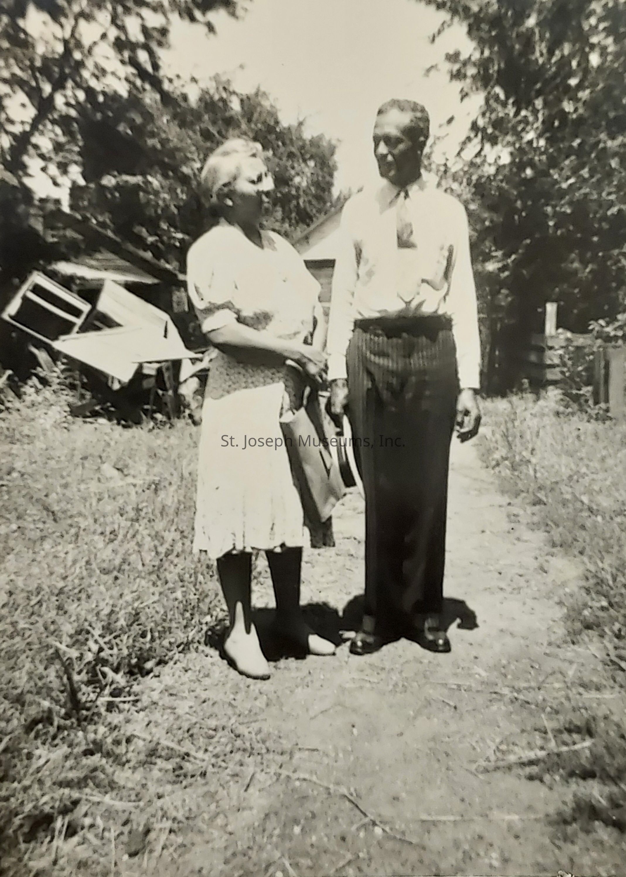 In the black-and-white photograph, an elderly couple stands side by side on a dirt pathway, surrounded by overgrown grass and trees, with the sun casting gentle shadows around them. The woman, dressed in a light-colored dress with soft patterns, holds a handbag, her feet slightly apart. She looks toward the man, who is wearing a white shirt and dark trousers, holding a hat in his hand. Behind them, there is a wooden structure, possibly a shed, tilted and partially hidden by foliage. The trees overhead create a dappled effect of light and shadow.