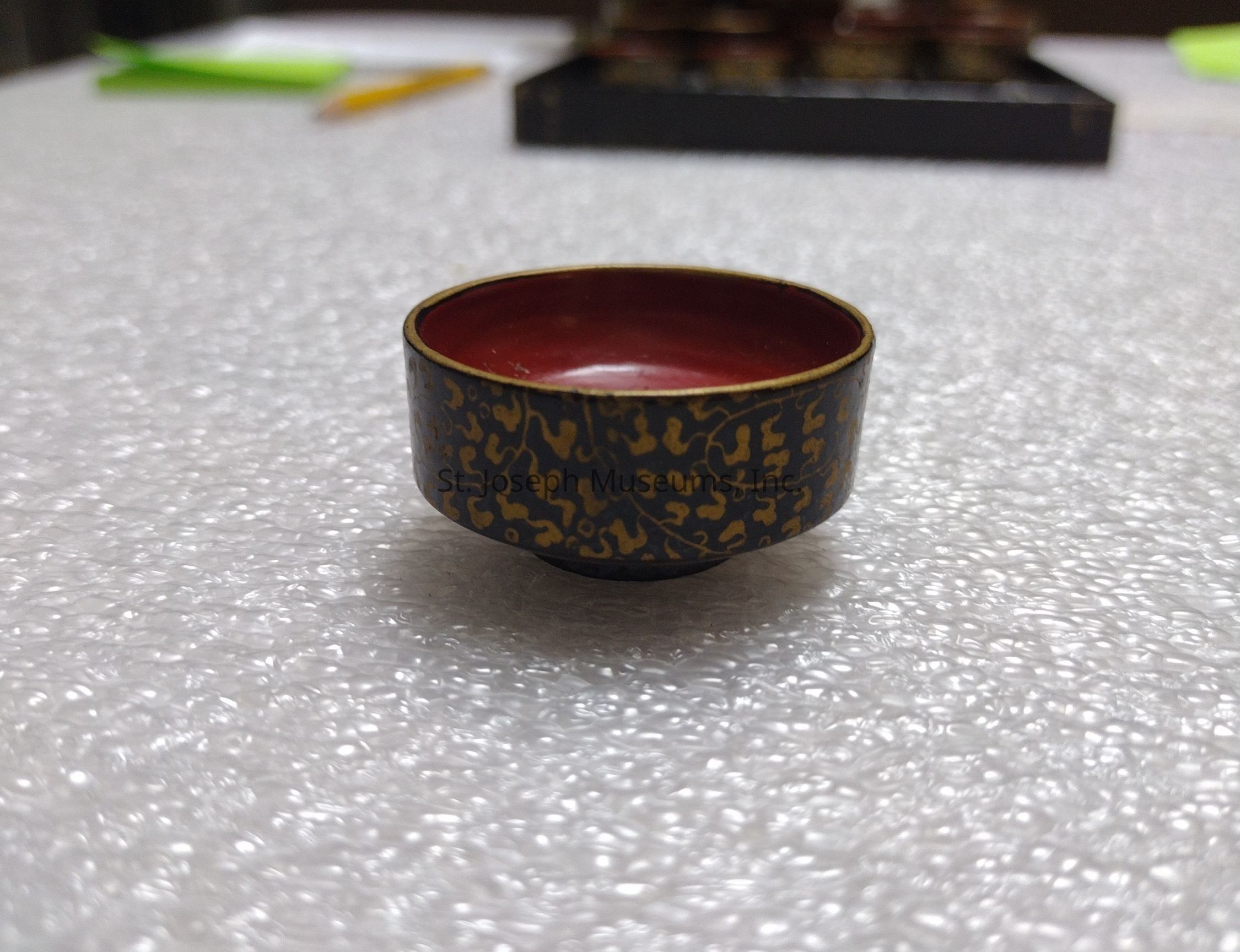 The image shows a small, intricately designed bowl placed on a textured surface. The bowl's exterior features a dark color embellished with delicate, gold patterning that appears to be of cultural or artistic significance. The interior is coated with a deep red finish, providing a striking contrast to its outer design. The patterned details on the bowl give it a refined and decorative appearance, suggesting it may be used for ceremonial or special occasions. In the background, a blurred tray or object can be seen, along with a pencil and a green paper, adding context to the setting as possibly being a workspace or an area focused on craft. The surface on which the bowl sits resembles bubble wrap, indicating the bowl might be in storage or being prepared for transport, suggesting care in handling due to the item's potential fragility or value. The composition directs focus primarily to the bowl, emphasizing its intricate details and craftsmanship while the softly lit environment highlights its textures and sheen.