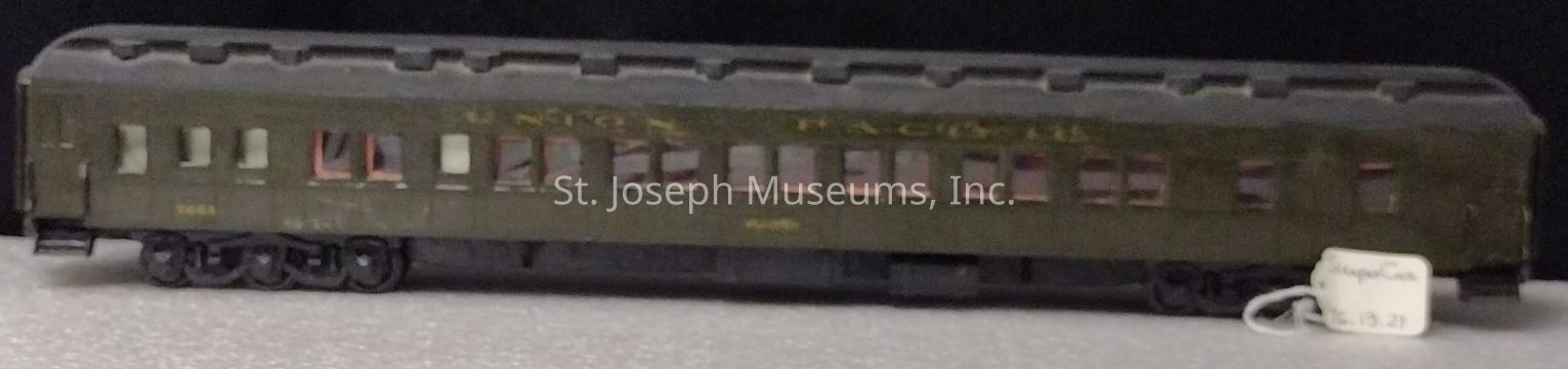 The image depicts a model of a vintage Union Pacific train car, characterized by its elongated rectangular shape and olive green color. The model includes numerous evenly spaced rectangular windows along the side, providing a glimpse inside, where miniature seats and details can be subtly discerned. Detailed elements like the roof’s distinct texture and the intricate design of the wheels showcase the craftsmanship behind the model. The words "Union Pacific" are subtly visible along the side, painted in a slightly faded shade of yellow. Attached to the car, there's a small white tag labeled "Baggage Car" and the number "$13.21," possibly indicating its classification and price. The surface beneath the model is a textured light gray, providing a neutral backdrop that highlights the train car’s historical design and color, while creating a nostalgic representation of freight transportation from a bygone era in meticulous small-scale form.