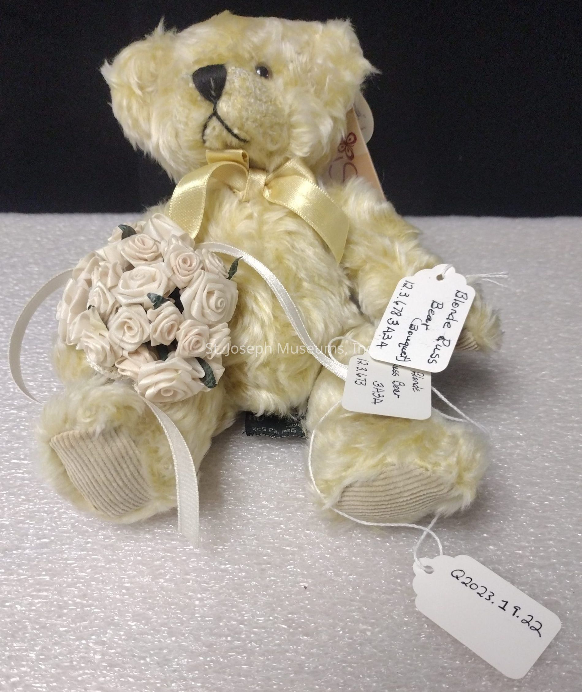 The image shows a plush teddy bear with soft, fluffy, light beige fur sitting upright against a gentle textured surface. The bear is adorned with a shiny, golden ribbon tied into a neat bow around its neck, adding a touch of elegance. In the bear's right paw, it delicately holds a small bouquet of cream-colored fabric roses, each detailed with subtle petals and accompanied by small green leaves, giving it a charming and romantic flair. Attached to the bear's left arm is a white tag with handwritten text, identifying it as a "Blonde Russ Bear" along with some alphanumeric details like "A213BA" and "QA2137." Another small, white tag dangles from the bear's right leg with a code "Q2023.18.22" handwritten on it, likely cataloging the bear as part of a collection or inventory. The stuffed animal embodies a sense of nostalgia and gifting, perfect for special occasions or as a collectible item.
