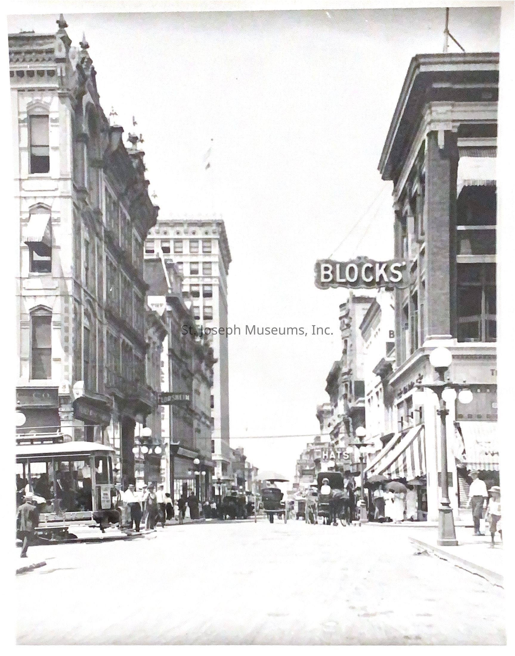 Black-and-white photo capturing a bustling street scene at the corner of 6th and Felix, looking west. Buildings with intricate architectural details line both sides, while a streetcar and pedestrians fill the scene. People dressed in period attire walk along the sidewalks as horse-drawn carriages move down the street.