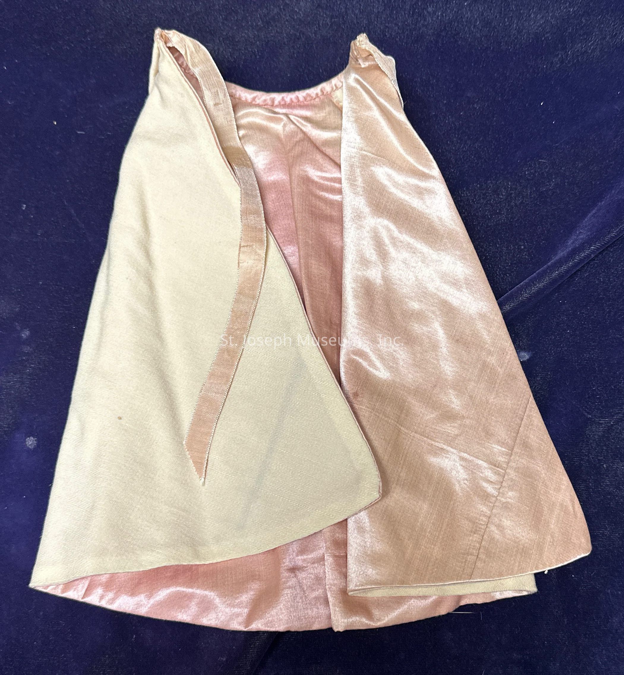 The image shows a garment, possibly a cape or an open-front jacket, with a smooth, shiny fabric, likely satin or silk, in a light beige or champagne color on the exterior, and a slightly darker pinkish hue on the interior. The garment is laid flat against a soft, dark blue background, which contrasts with the lighter tones of the fabric. The edges of the garment are meticulously finished, creating clean lines that highlight the smooth texture of the material. The inner panel features a subtle seam detail that provides a glimpse into the garment's construction. The lighting in the image emphasizes the sheen on the fabric, creating a glossy effect that suggests a luxurious quality. The simple yet elegant design implies it could be used as a formal accessory, adding a touch of sophistication to an outfit. The overall presentation is neat, allowing the garment's texture and color to be the focal points.