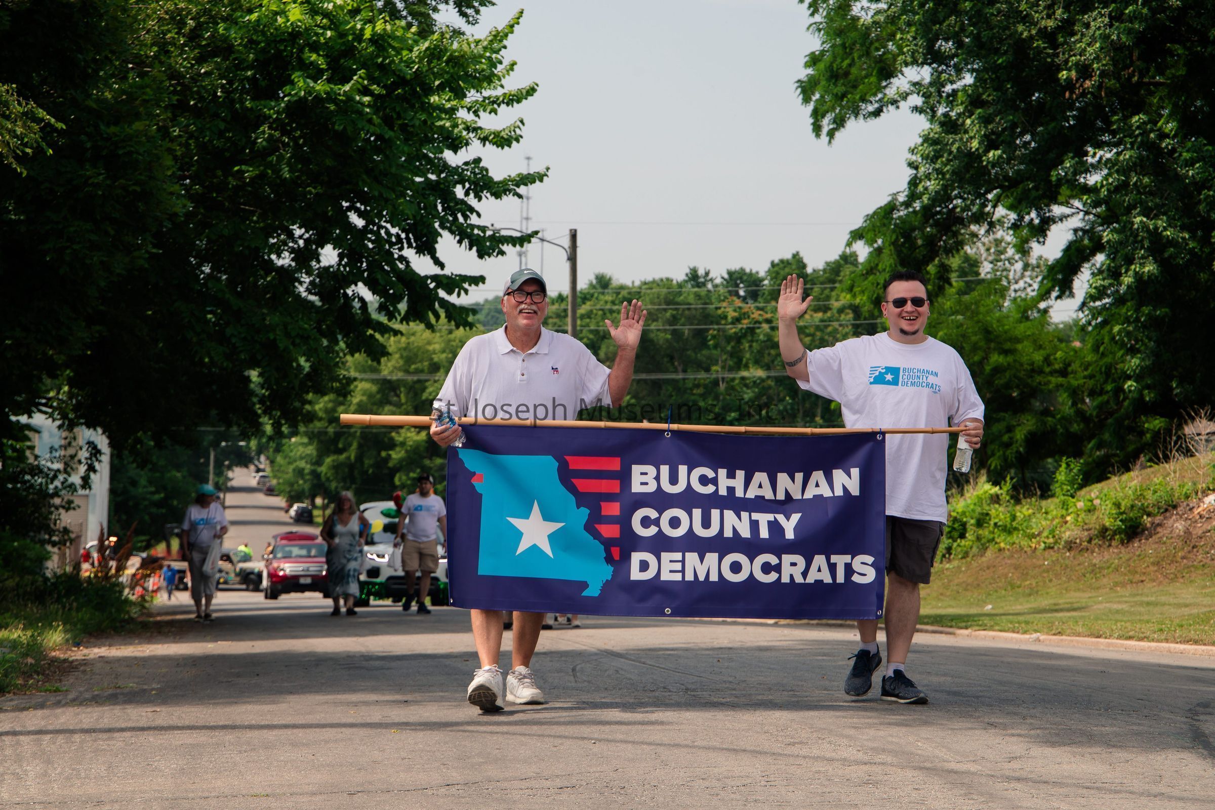 In the image, two men are walking down a tree-lined street while holding a large banner that reads “Buchanan County Democrats,” featuring a blue background with a white star and a red-striped design. The man on the left is wearing a white polo shirt, a cap, and holding a water bottle, smiling and waving at the camera. The man on the right is wearing a white T-shirt with matching design elements, also smiling and waving, holding a water bottle as well. Behind them, more people can be seen walking, wearing similar attire, participating in what appears to be a community parade. The background is lush and green, with trees lining both sides of the street, and several vehicles are visible further down the road, enhancing the sense of a vibrant and active event.