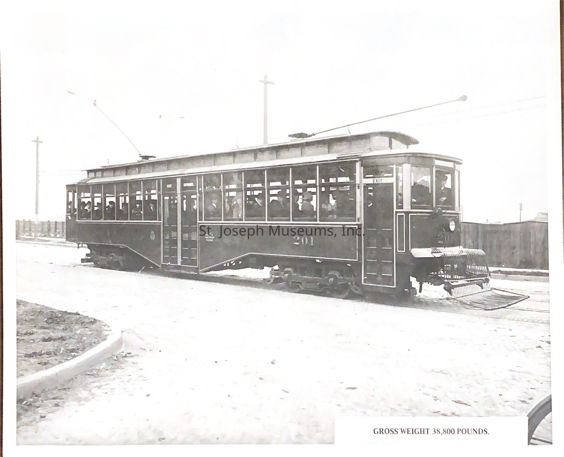 Black-and-white photograph of a streetcar, labeled with the number 201, filled with passengers and set on tracks running through an urban area, showcasing a design typical of early 20th-century public transportation. The car features an ornate frontal design with rounded edges and multiple windows, reflecting its ability to accommodate numerous riders.