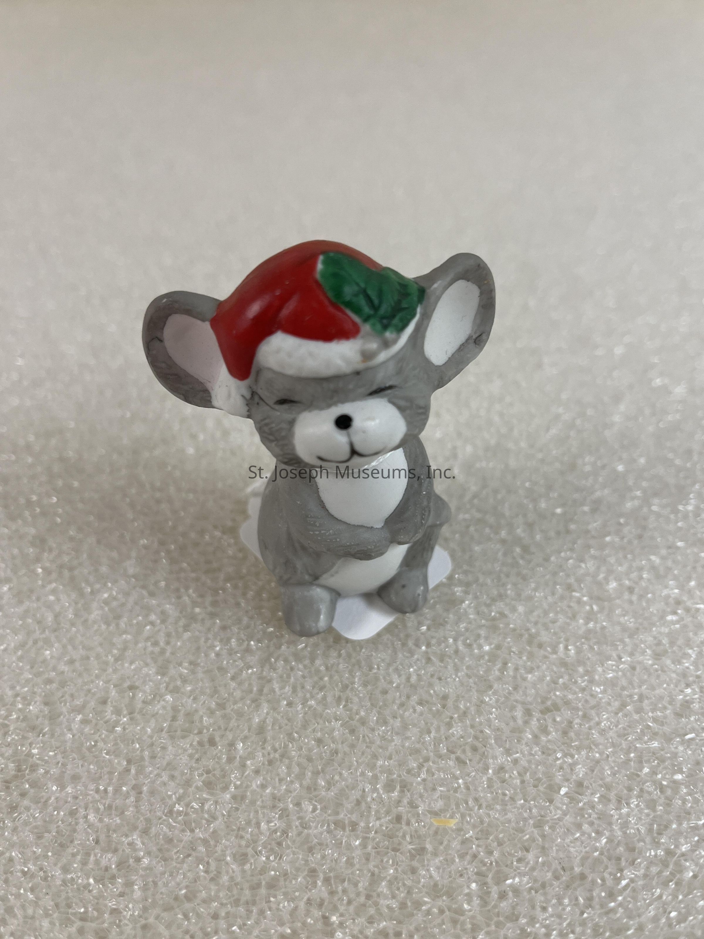 In the image, there is a small figurine of a gray mouse standing on a textured surface. The mouse is wearing a red Santa hat with a green leaf attached to it, and it has its eyes closed, creating an expression that appears content or happy. Its body is primarily gray, with white details on its stomach and face, giving it a soft, inviting look. The ears are large and rounded, contributing to its charming appearance. The overall effect of the figurine is festive, bringing to mind holiday cheer and the spirit of Christmas. The simplicity of the background enhances the focus on the mouse, highlighting its detailed features and the vibrant colors of its hat. The textured surface adds a subtle, pleasing complexity to the setting, engaging the viewer without distracting from the whimsical charm of the figurine. The posture of the mouse, along with its festive attire, suggests an air of warmth and celebration, making it a delightful visual representation of holiday themes.