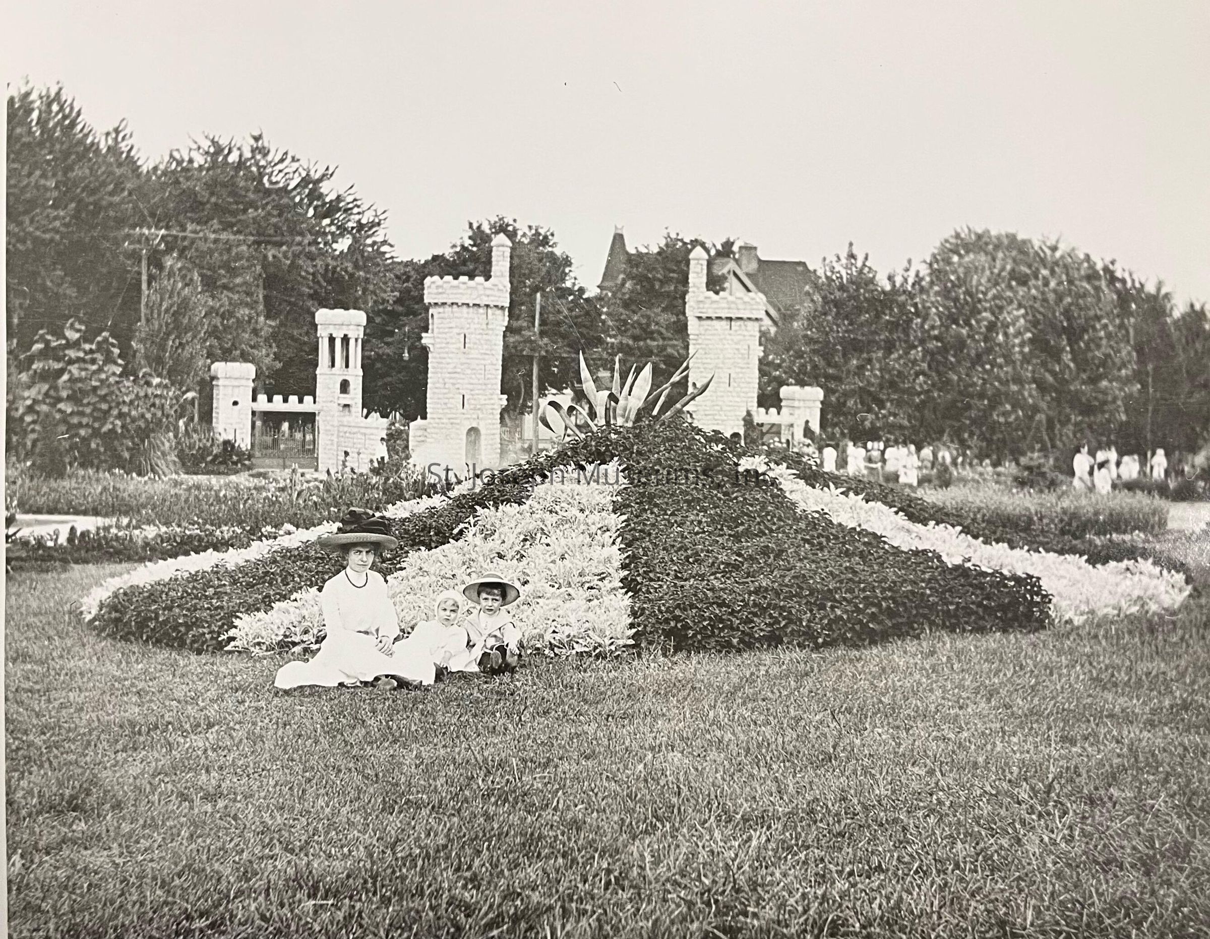 Black-and-white photograph of the Krug Park Flower Beds, in front of which a woman and two children sit on the grass in front of an ornately designed bed of flowers shaped in a geometric pattern. Behind them, stone castle-like structures rise amid lush trees, adding an enchanting backdrop to the scene.