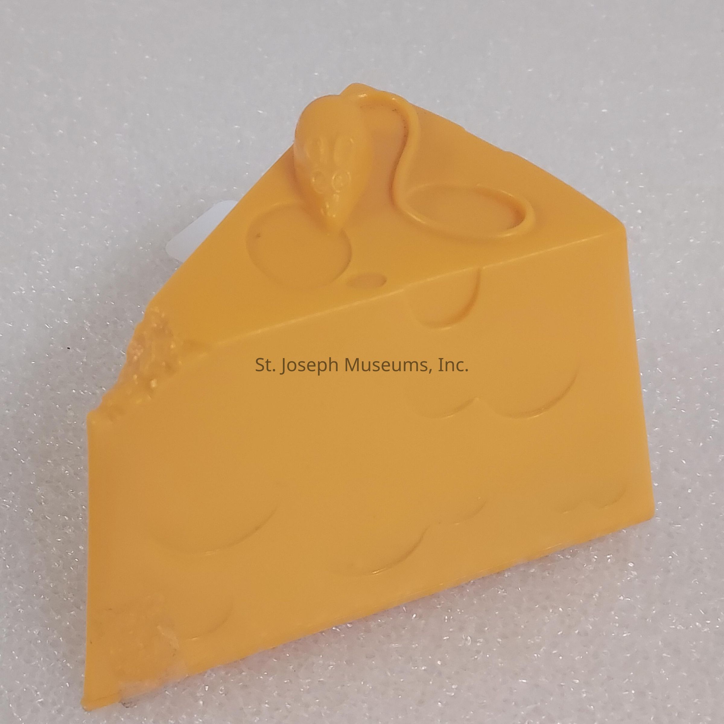 The image depicts a realistic-looking piece of yellow cheese, likely made of plastic or another synthetic material, placed on a textured white surface. The cheese is wedge-shaped and features cartoon-like holes on its sides, mimicking the appearance of traditional Swiss cheese. Atop the wedge is an adorable small depiction of a mouse, complete with rounded ears, tiny eyes, and a long tail that curls back onto the cheese surface, adding a whimsical and playful touch to the scene. The overall presentation is charming and suggests a humorous take on the classic pairing of mice and cheese, making it an appealing decorative or novelty item.