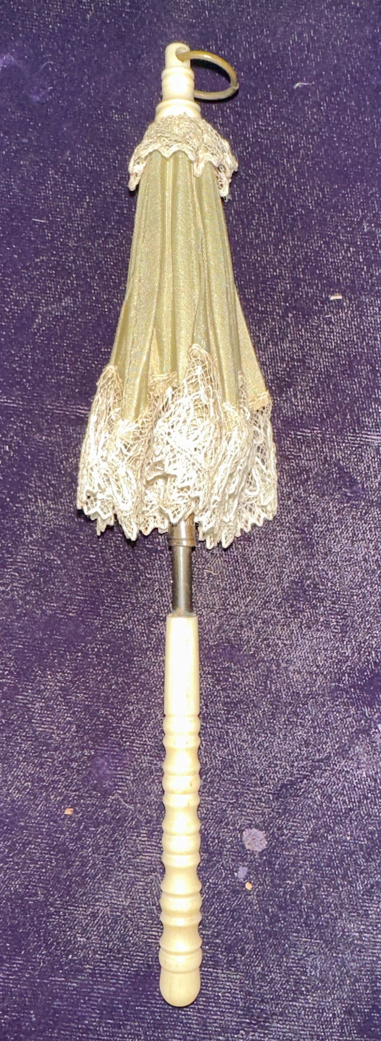 The image depicts a vintage parasol closed against a deep purple textured background. The parasol features an ornate design, characterized by light beige fabric adorned with intricate lace trim along the edge, creating an elegant and antique appearance. The handle is carved from a material resembling ivory, exhibiting a series of decorative rings and a smooth, polished finish. At the top of the parasol, there is a loop, suggesting it could be easily carried or hung. The overall look of the parasol hints at a bygone era, reflecting a classic style often associated with historical fashion. Its delicate details and craftsmanship suggest it was likely intended for decorative purposes or gentle sun protection rather than practical use against rain. This combination of elements creates an impression of sophistication and timeless charm, capturing an essence of elegance typically valued in historical accessories.