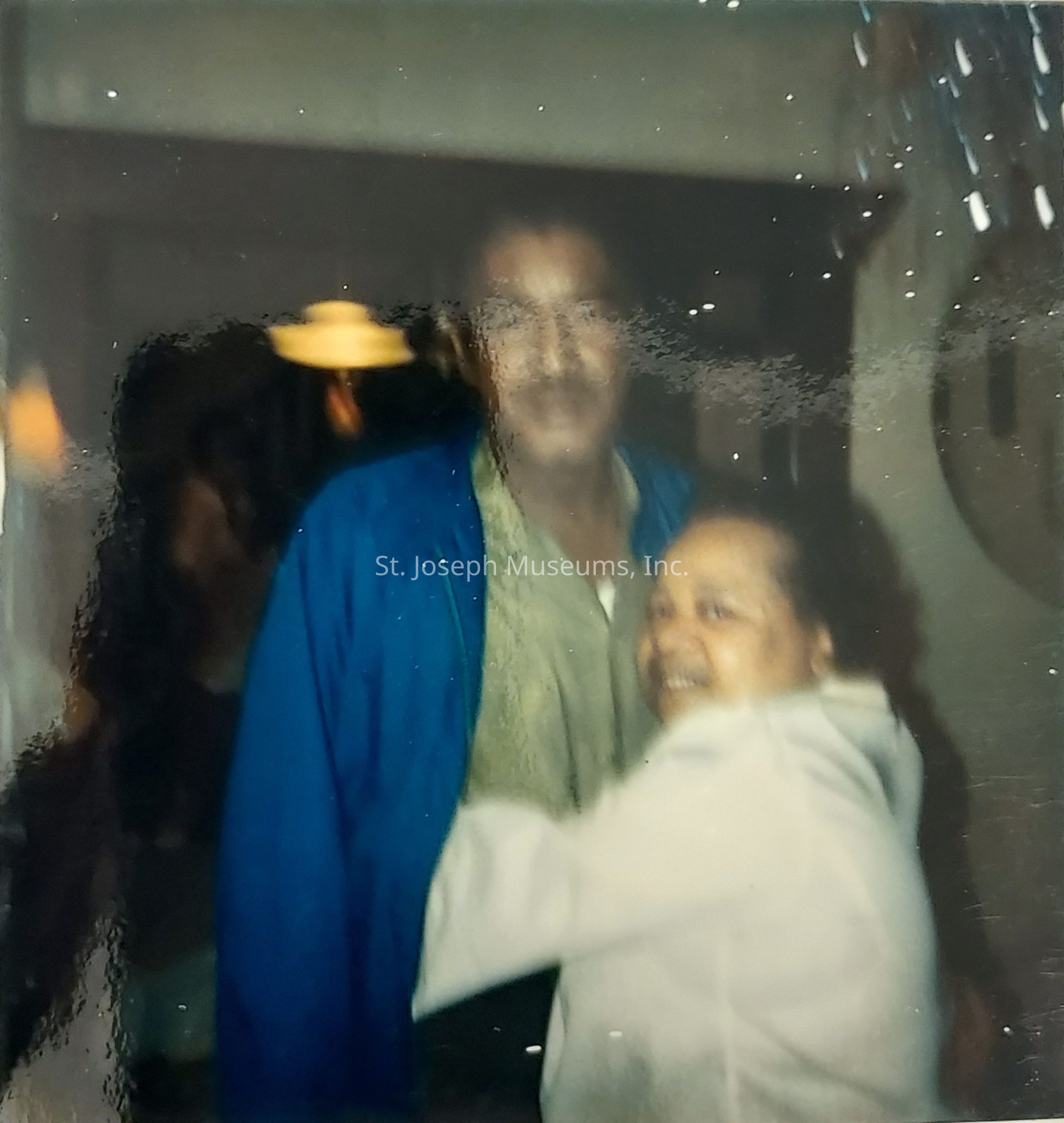 In the image, a man and a woman are embracing indoors, with the man wearing a blue jacket over a beige shirt and the woman dressed in a light-colored top. The setting appears to be warmly lit, adding a cozy ambiance, and there are blurred background elements, possibly indicating other people or objects, which suggest this could be a casual or family gathering. The overall atmosphere of the photo conveys a sense of warmth and familiarity, capturing a moment of connection and affection between the two individuals as they smile, creating a timeless and candid snapshot of a shared moment.