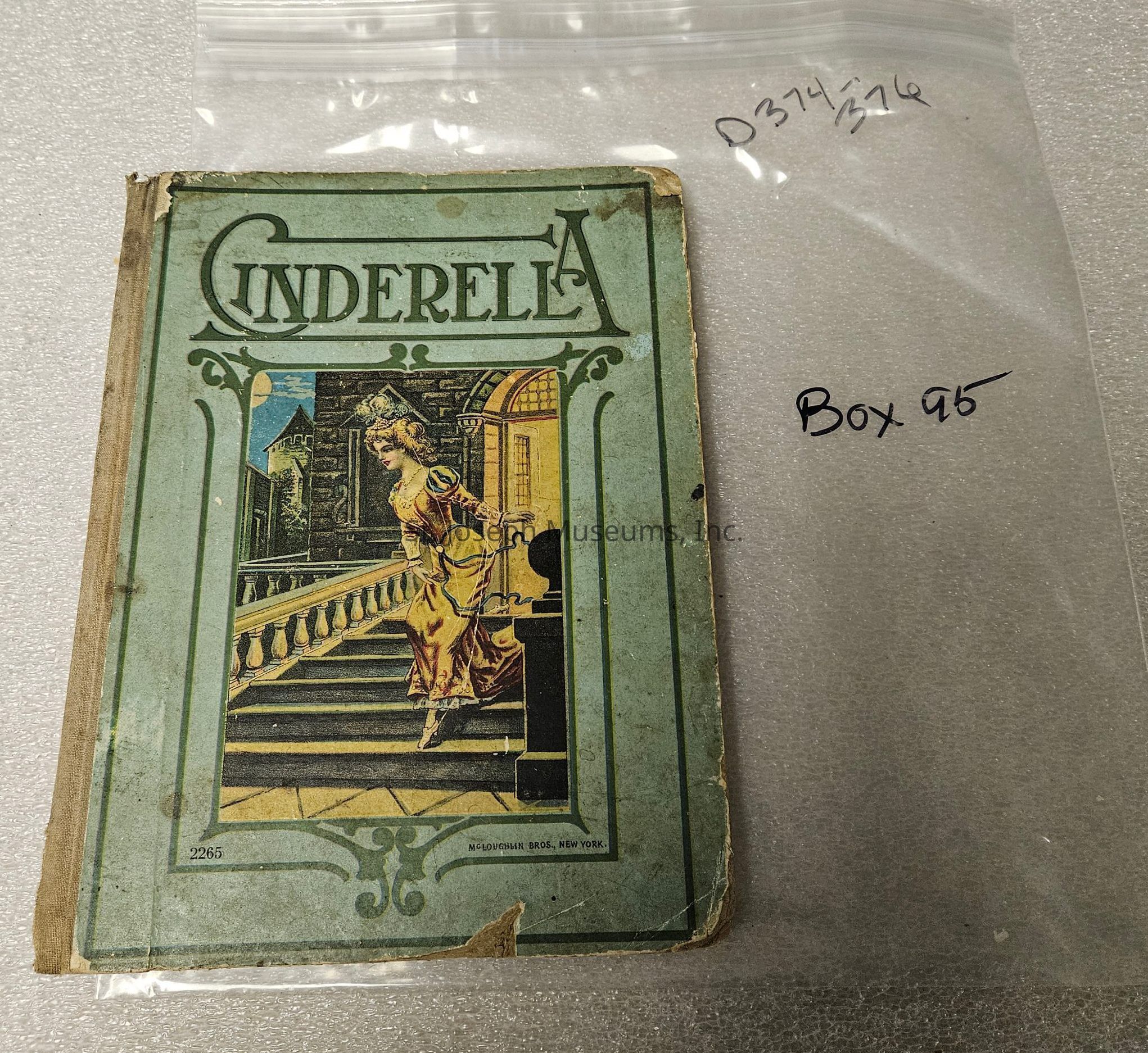 The image shows an old, worn copy of a book titled "Cinderella," with a green and slightly tattered cover, featuring an illustration of Cinderella in a yellow dress, descending a grand staircase in a palace setting, with a castle visible in the background; the book is placed next to a clear plastic bag labeled "Box 95" and "D374/376," which suggests archival storage or cataloging, and the book's publisher is noted as McLoughlin Bros., New York, indicating its historical value and possibly vintage publication.
