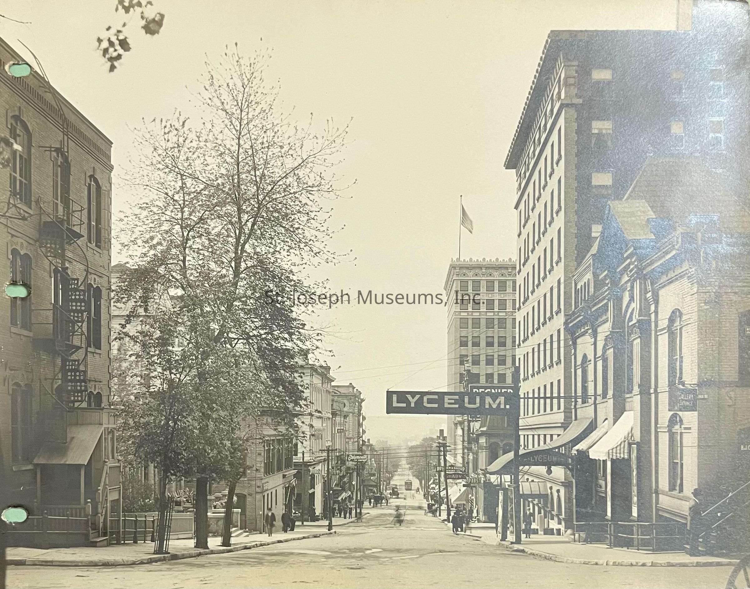 Black-and-white photograph capturing a historic street view looking south on 4th Street towards Francis, highlighting the exterior of the Lyceum Theater. The scene features vintage architecture with brick buildings lining the streets, some adorned with awnings and ornate detailing. A few pedestrians can be seen strolling down the sidewalk.