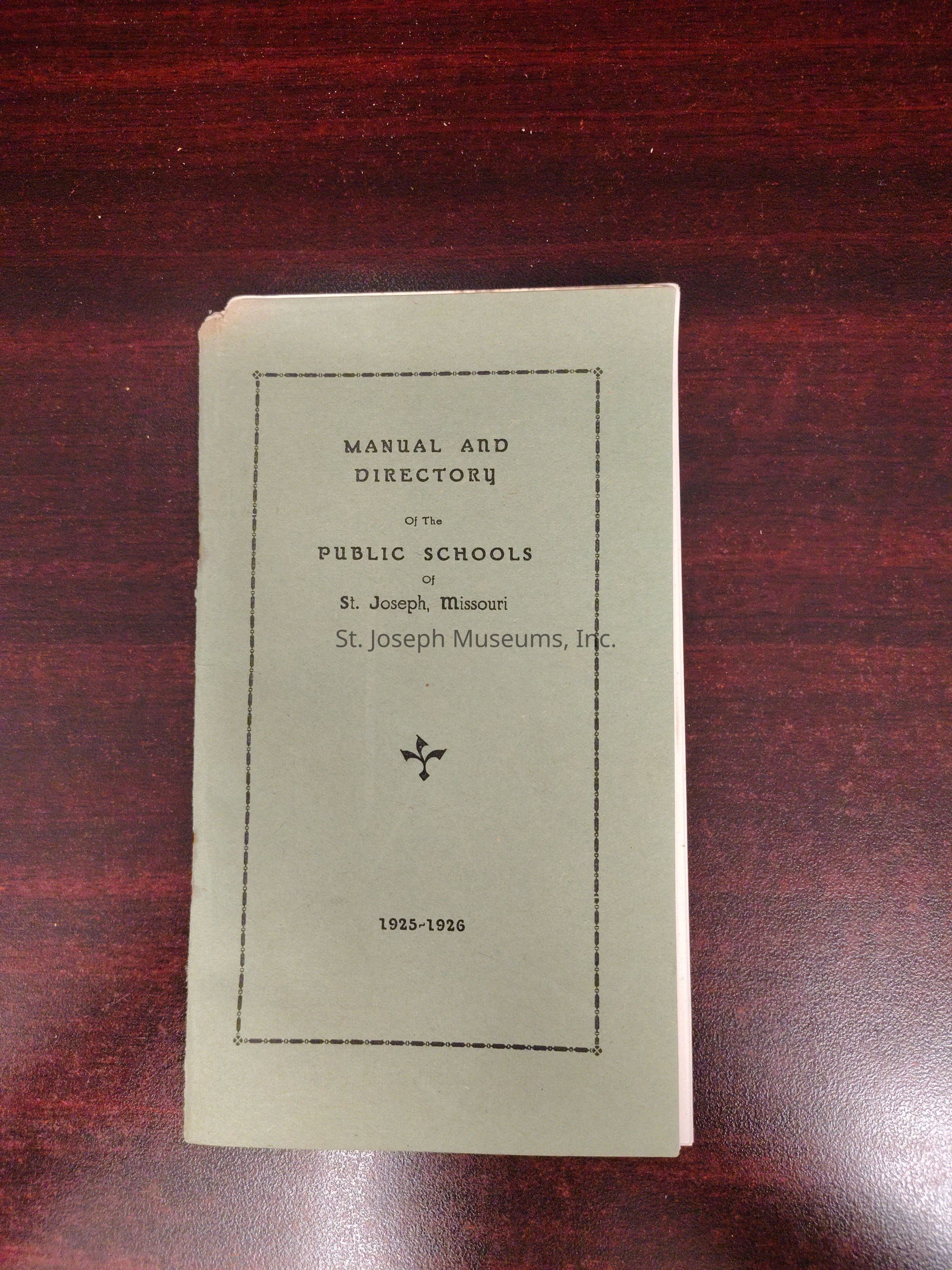 The image features a vintage booklet with a greenish cover on a dark wood surface, titled "Manual and Directory of the Public Schools of St. Joseph, Missouri," for the year 1925-1926. The cover design is simple, with a rectangular border surrounding the text, as well as a small decorative symbol positioned near the bottom of the cover. The edges of the booklet show slight wear, indicating its age and usage over the years. The carefully aligned text, printed in black ink, contrasts against the muted background of the cover, imparting an understated but classic appearance typical of early 20th-century printed materials.