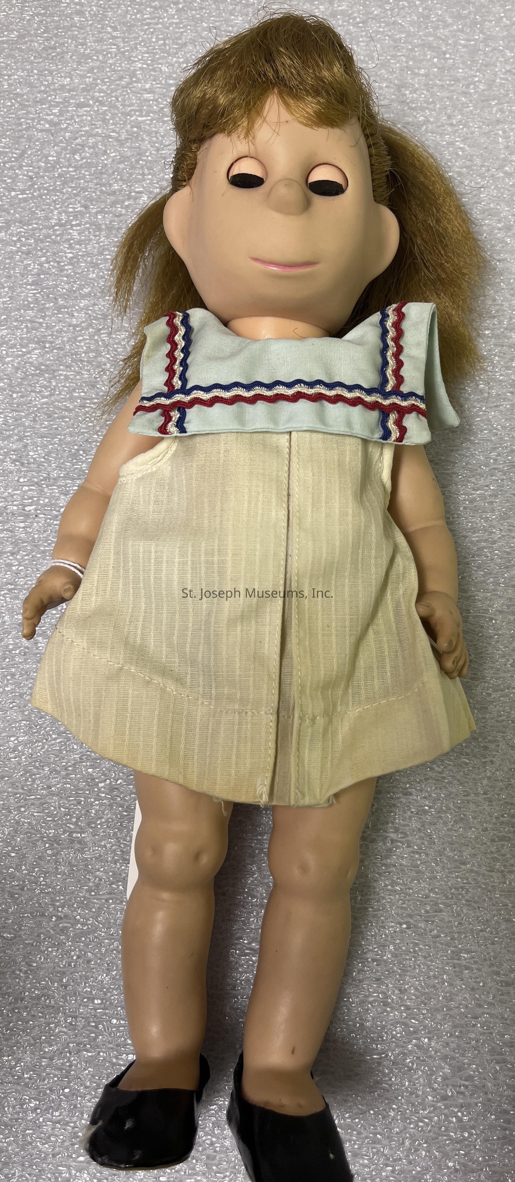 The image depicts a vintage doll with a unique and slightly unusual design, featuring a soft cloth body and a rubber face. The doll has round, black eyes without visible pupils and a small, subtle smile. It is dressed in a light-colored, sleeveless dress with a collar adorned with blue and red trim, adding a touch of color and contrast to the outfit. The hair is light brown and somewhat messy, suggesting years of handling and play. The doll's plastic legs are smooth and slightly bent at the knees, and it wears black shoes, completing its quaint, old-fashioned appearance. The entire image is set against a plain, textured background, likely enhancing the presentation of the doll by focusing attention solely on its features and attire.