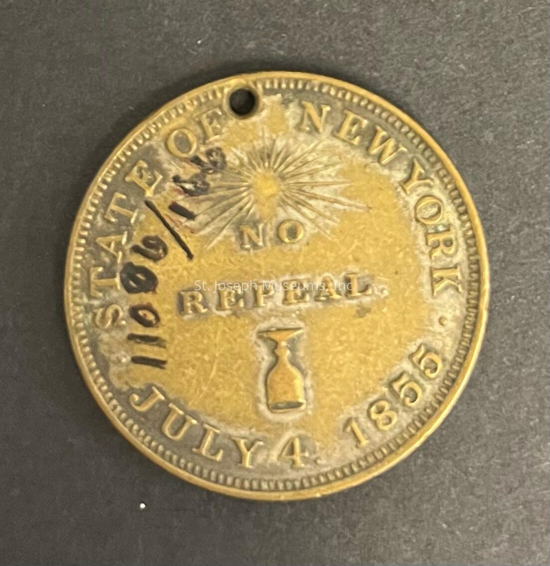 The image depicts an old, round brass token with a stamped design related to the state of New York. The center features the words "NO REPEAL" with an hourglass symbol beneath it, emphasizing a statement of permanence or protest against change. Surrounding the central message, the inscription reads "STATE OF NEW YORK" along the top edge and "JULY 4, 1855" along the bottom, indicating the date, likely highlighting an event of historical significance. A radiating sun emblem appears above "NO," and there is a small hole near the top, possibly for hanging or attaching the token.