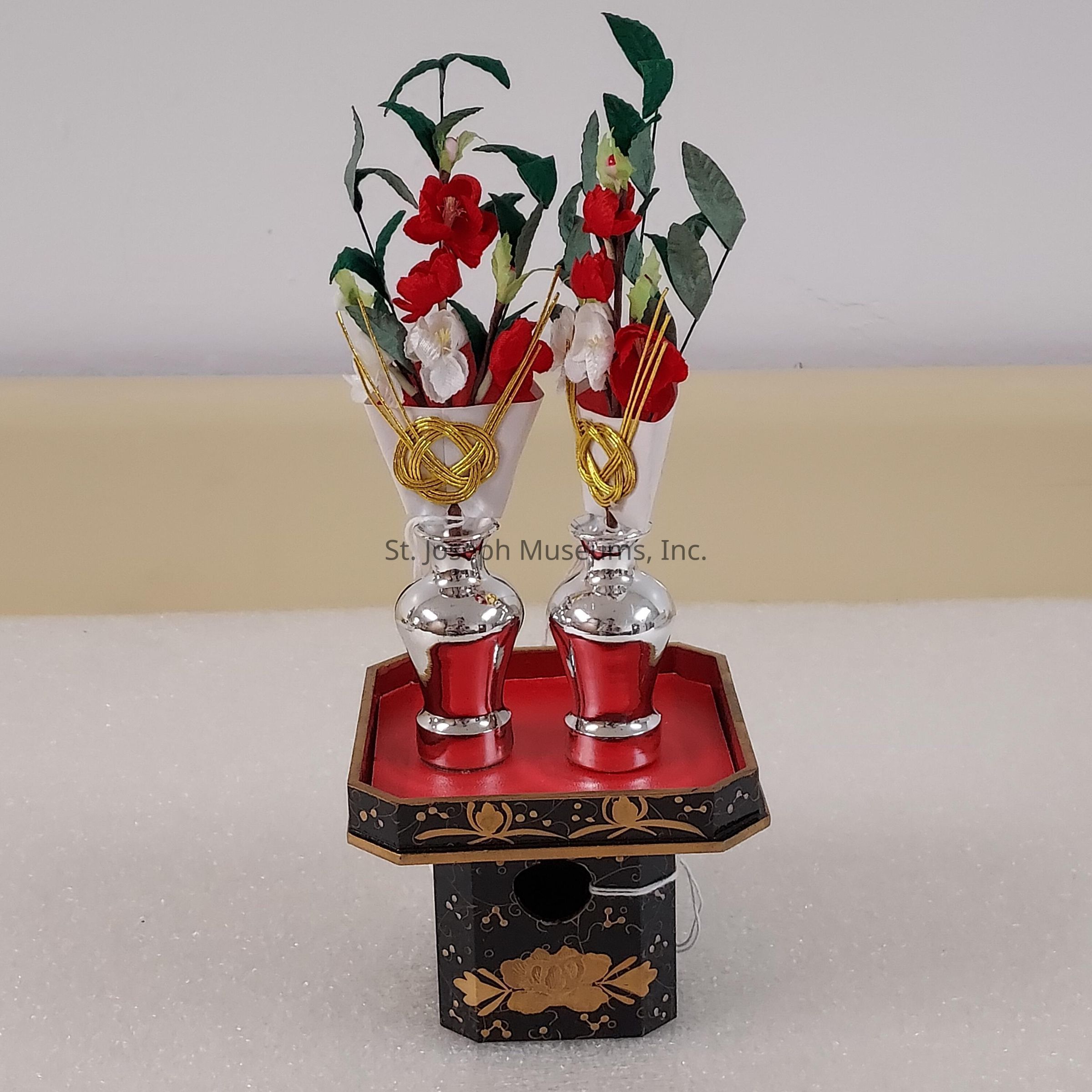 The image depicts an intricately designed arrangement featuring two vases with metallic bases, each adorned with delicate, artfully crafted red and white flowers accompanied by green leaves. The vases are elegantly positioned on an ornate, black lacquered stand embellished with gold floral patterns and details. The decorative golden cords wrapped around the floral stems add a touch of sophistication to the arrangement. The stand has a red top surface, perfectly complementing the vivid colors of the flowers and creating a harmonious visual balance. This arrangement is set against a neutral background, emphasizing its artistic and cultural significance.