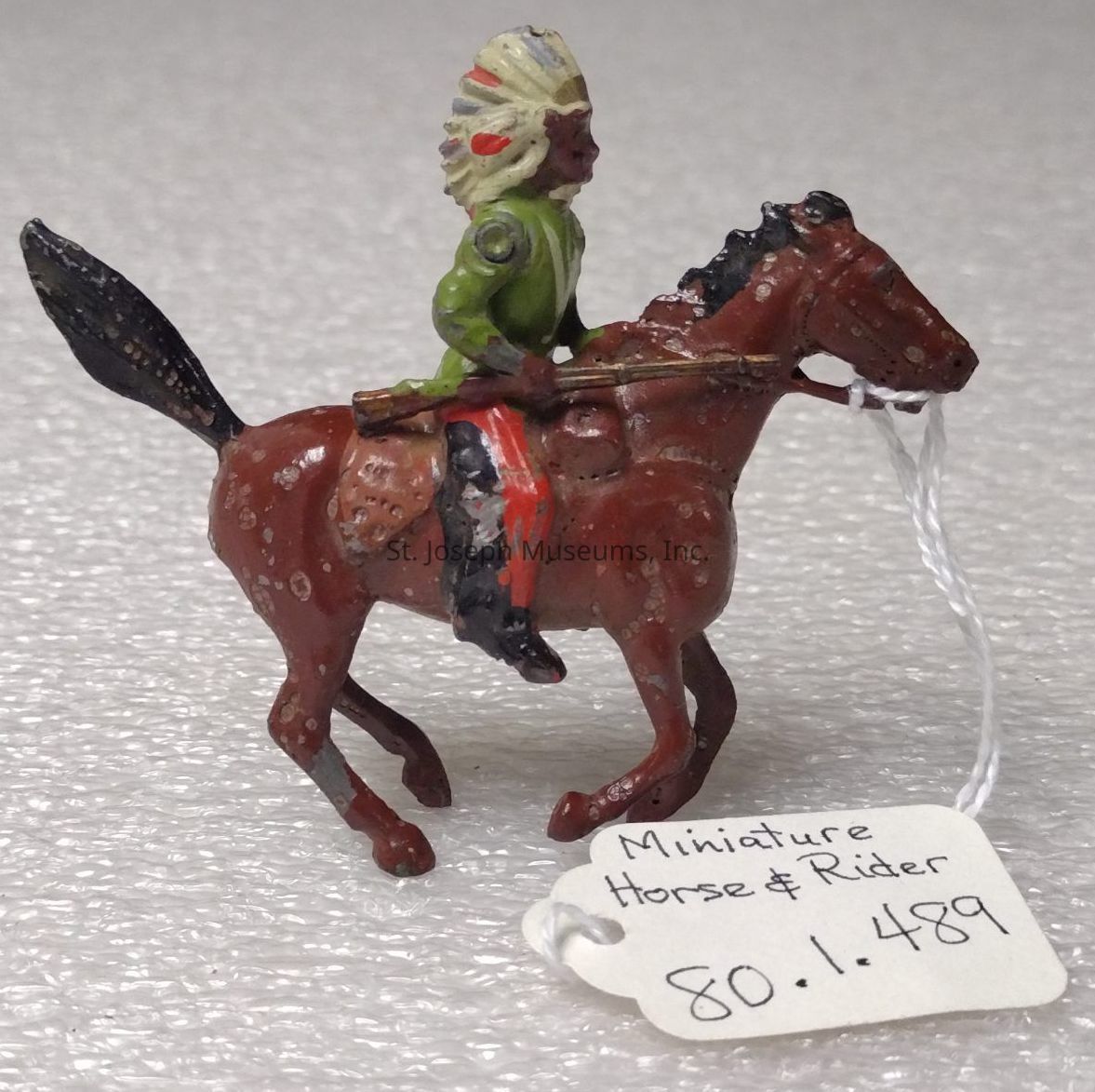 The image features a small painted figurine depicting a rider on a horse. The rider is wearing a green outfit and a headdress with feathers in shades of red and white. The horse is brown with red and black detailing, suggesting a dynamic motion. The figurine is crafted with intricate details, highlighting the expressive stance of both the rider and horse. They are set against a neutral background, emphasizing the vivid colors and texture of the model. A white string is attached to the horse's mouth, perhaps simulating reins. Additionally, there is a tag with handwritten text that reads "Miniature Horse & Rider" followed by the numbers "80.1.489," suggesting it might be part of a collection or cataloged item. The overall composition of the figurine, with its detailed craftsmanship, evokes a sense of movement and cultural significance, capturing the essence of traditional attire and the bond between rider and horse in a miniature form.