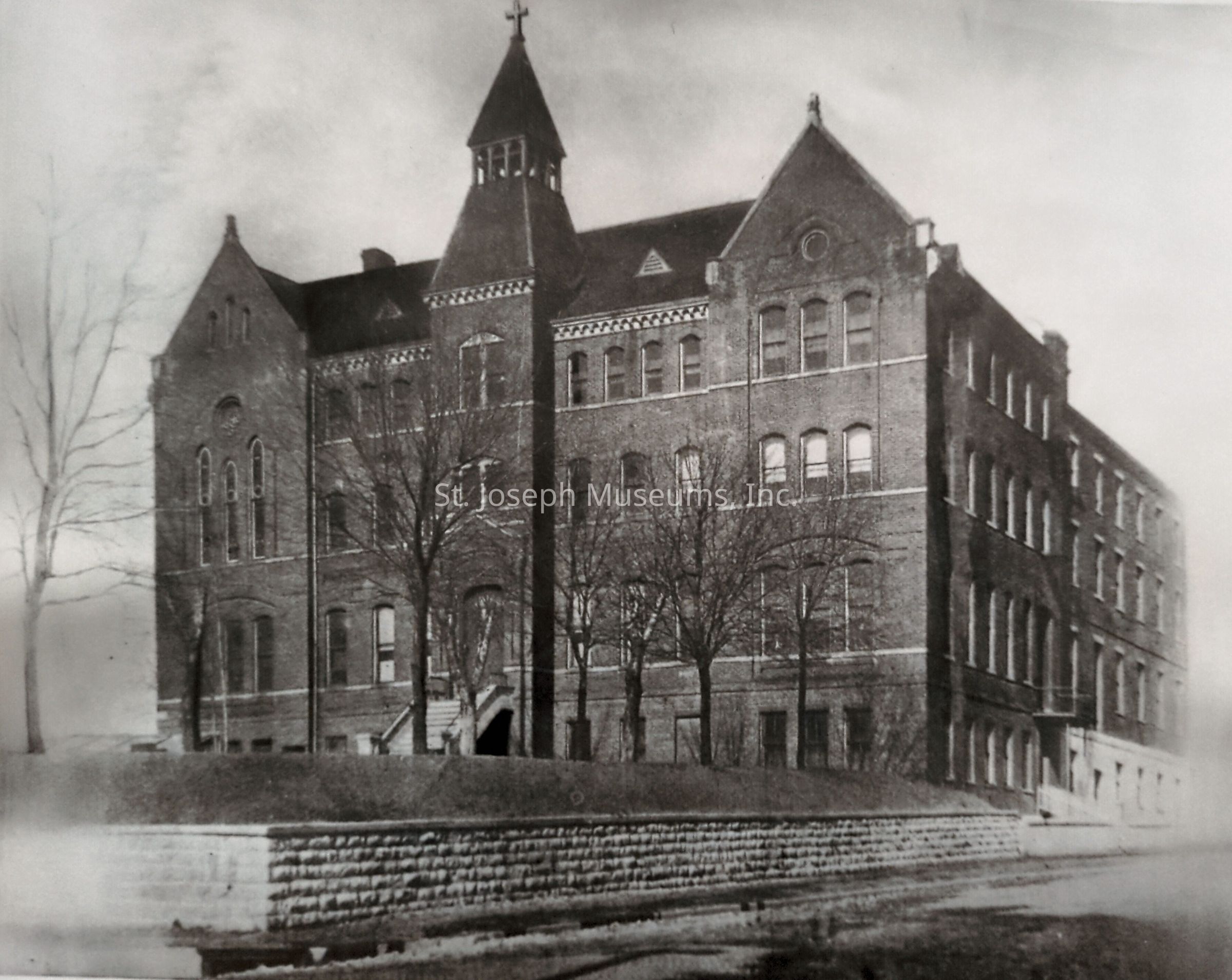 Black-and-white image depicting the exterior of St. Joseph Commercial College at 13th and Henry Streets, a stately brick building built in 1858 by the Christian Brothers. Its architecture features multiple stories, a pointed roof, and arched windows. The solid structure is complemented by bare trees in the foreground.