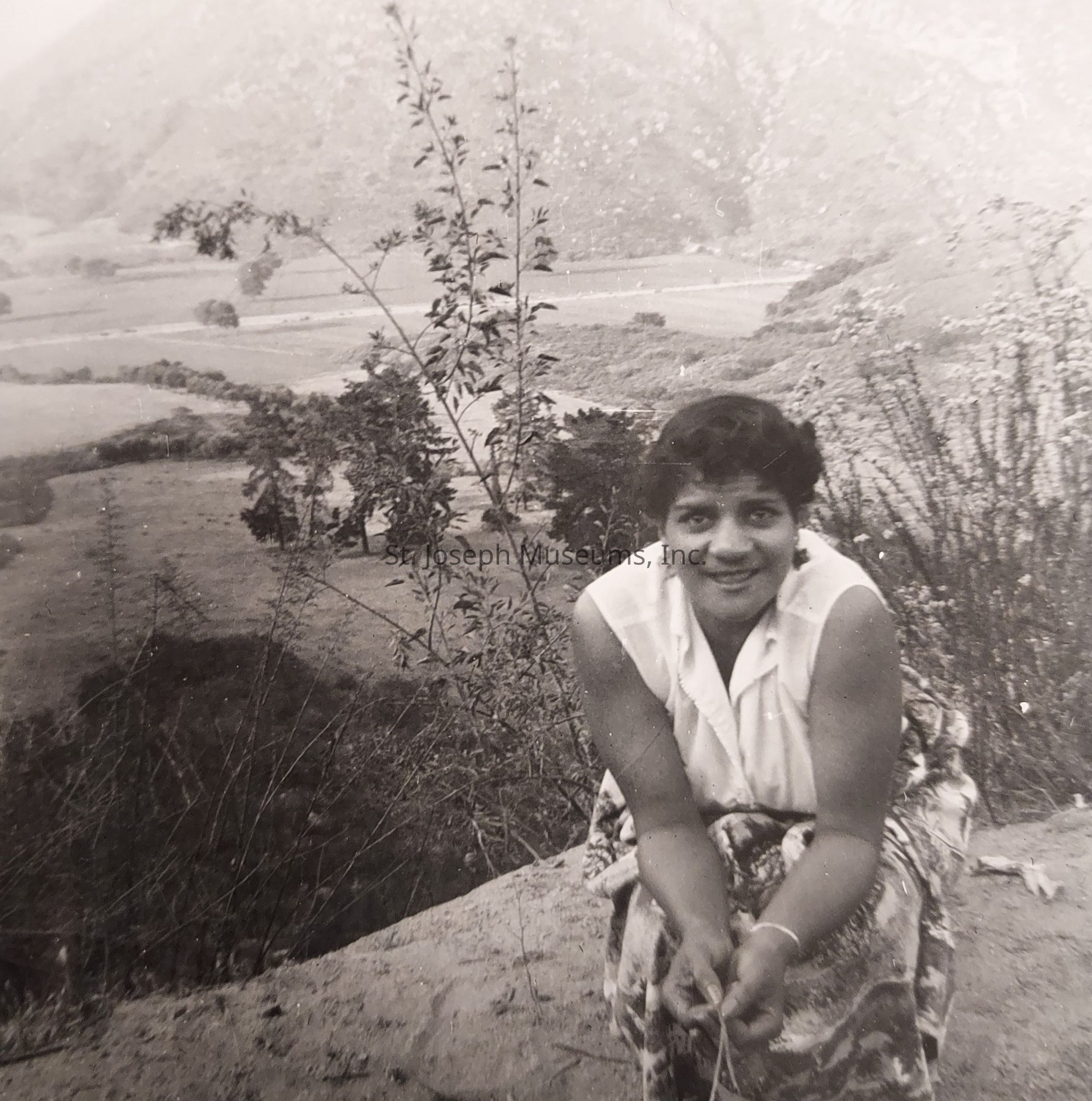 In this black and white photograph, a woman is kneeling and smiling warmly towards the camera, creating an inviting atmosphere. She is wearing a sleeveless blouse and a patterned skirt, appearing relaxed and content. The setting around her is natural and serene, with varied vegetation including tall, slender plants in the foreground, adding a touch of wildness to the scene. The backdrop reveals expansive, rolling hills and vast open spaces, suggesting a location that is both rural and picturesque. The contrast between the woman’s cheerful expression and the tranquility of the environment captures a moment of harmony with nature. The soft hills in the distance create a sense of depth, drawing the viewer’s eye further into the landscape. This image, devoid of color, relies on shades and textures to convey emotion and atmosphere, enhanced by the woman's engaging demeanor and the surrounding scenic beauty, making it an evocative snapshot of a simple yet profound moment in time.