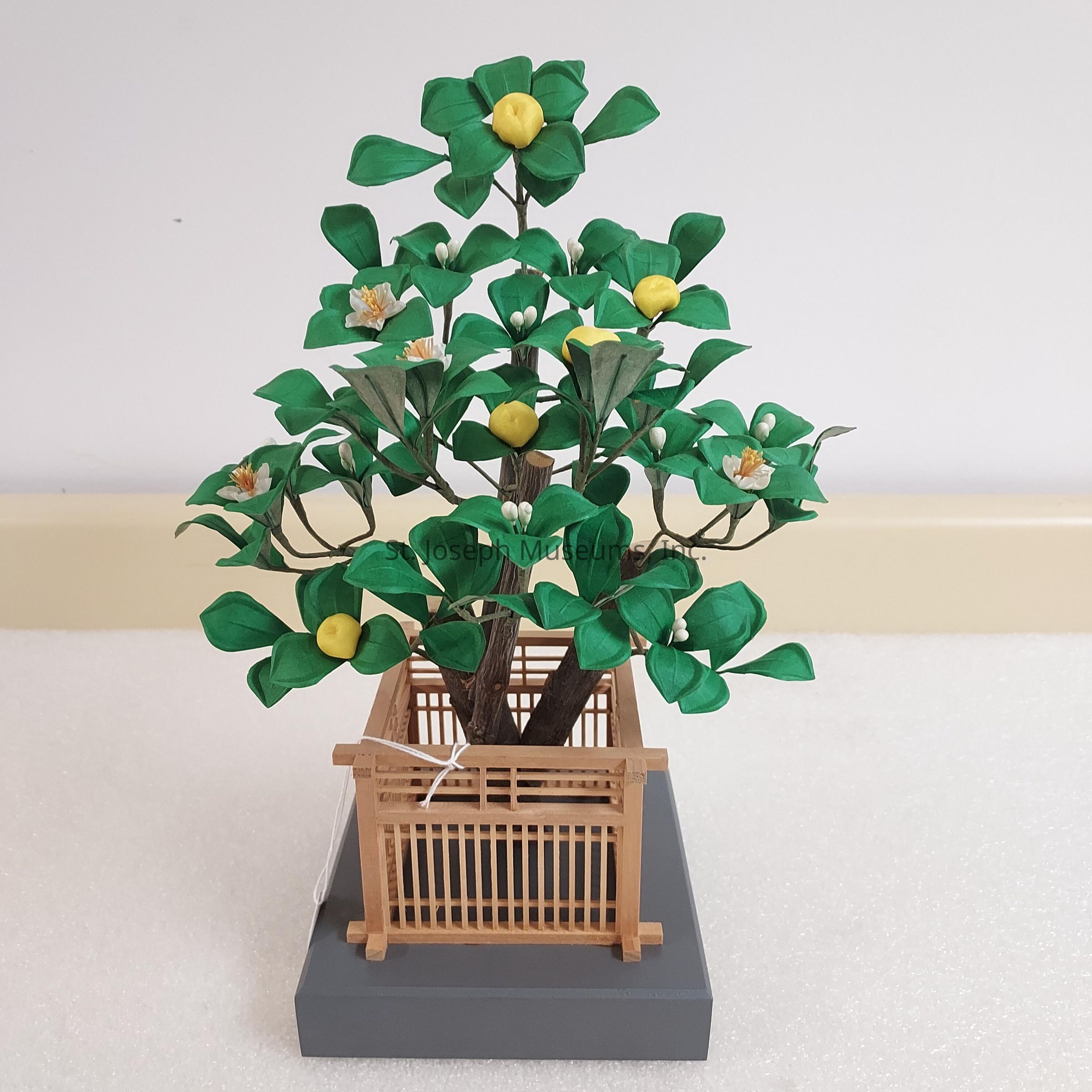 The image shows a delicate, handcrafted model of a tree, featuring bright green leaves made from paper or a similar material and small, round, yellow fruits, along with a few white blossoms, all arranged intricately on thin branches. This miniature tree is placed in a small, wooden lattice crate on top of a gray platform, with a clean white background and a cream-colored strip along the lower edge, enhancing the visual simplicity and focus on the tree itself. Each detail contributes to the overall impression of natural beauty, showcasing the skill and patience involved in its creation.
