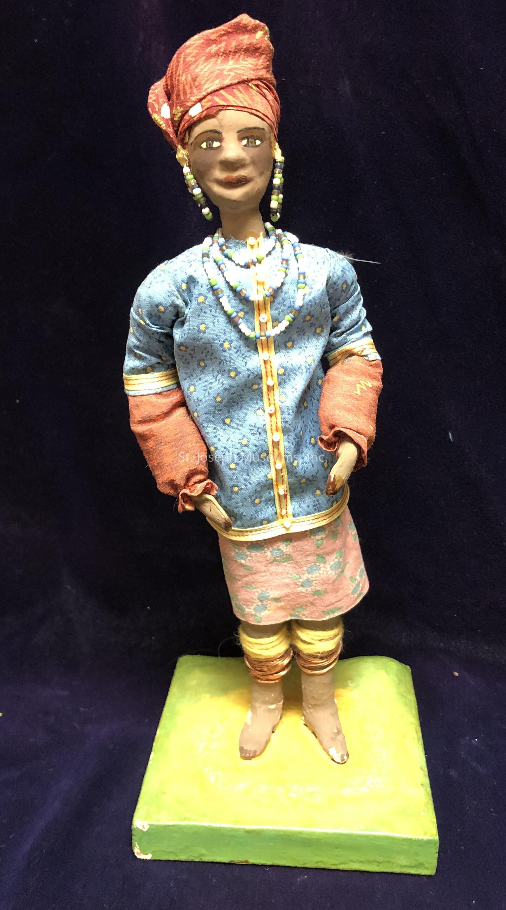 The image features a handcrafted doll standing on a square green base against a dark blue background. The doll is adorned in vibrant and traditional attire, including a blue long-sleeved top with a yellow floral pattern and contrasting yellow accents along the front and sleeves. Layered beneath is a peach-colored skirt decorated with a subtle blue floral design, complementing the top. The figure is accessorized with multiple beaded necklaces and earrings in hues of blue, white, and green. The doll’s head is wrapped in a striking red headpiece with subtle patterns, matching the red fabric wrapped around its arms. The legs are wrapped in yellow and brown, simulating traditional leg adornments, and the feet are shaped to appear bare, showcasing attention to detail in its craftsmanship. The doll’s features are intricately detailed, with painted facial elements conveying a serene expression, capturing a sense of cultural richness and artistic authenticity.