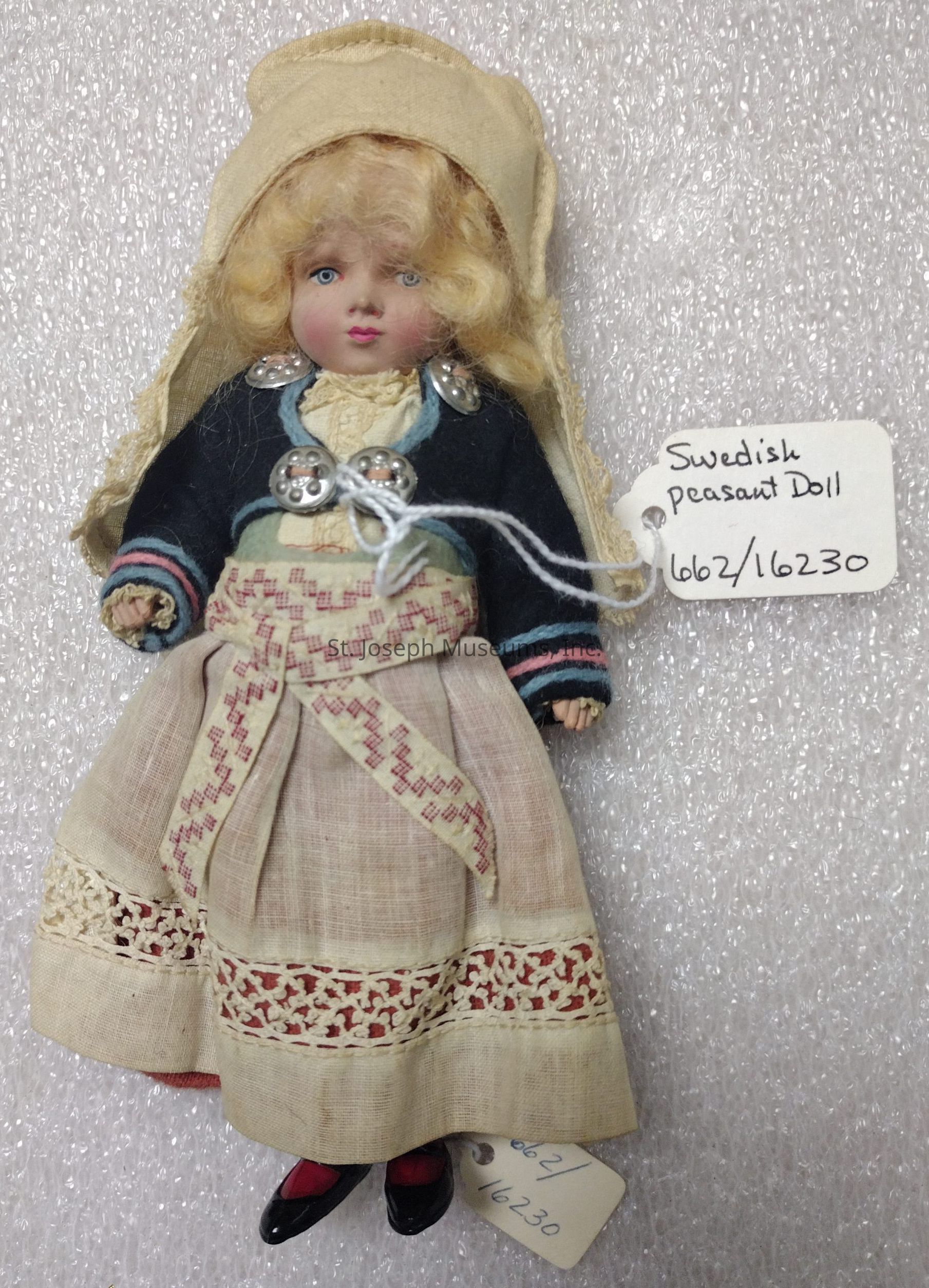 The image features a Swedish peasant doll dressed in traditional attire, with a cream-colored headscarf framing its curly blonde hair. The doll dons a dark blue jacket adorned with silver buttons over a cream blouse. A decorative apron, embellished with red embroidery and lace trim, wraps around the skirt. The delicate details of the clothing, including the intricate stitching and textures, highlight the craftsmanship involved in creating this piece.