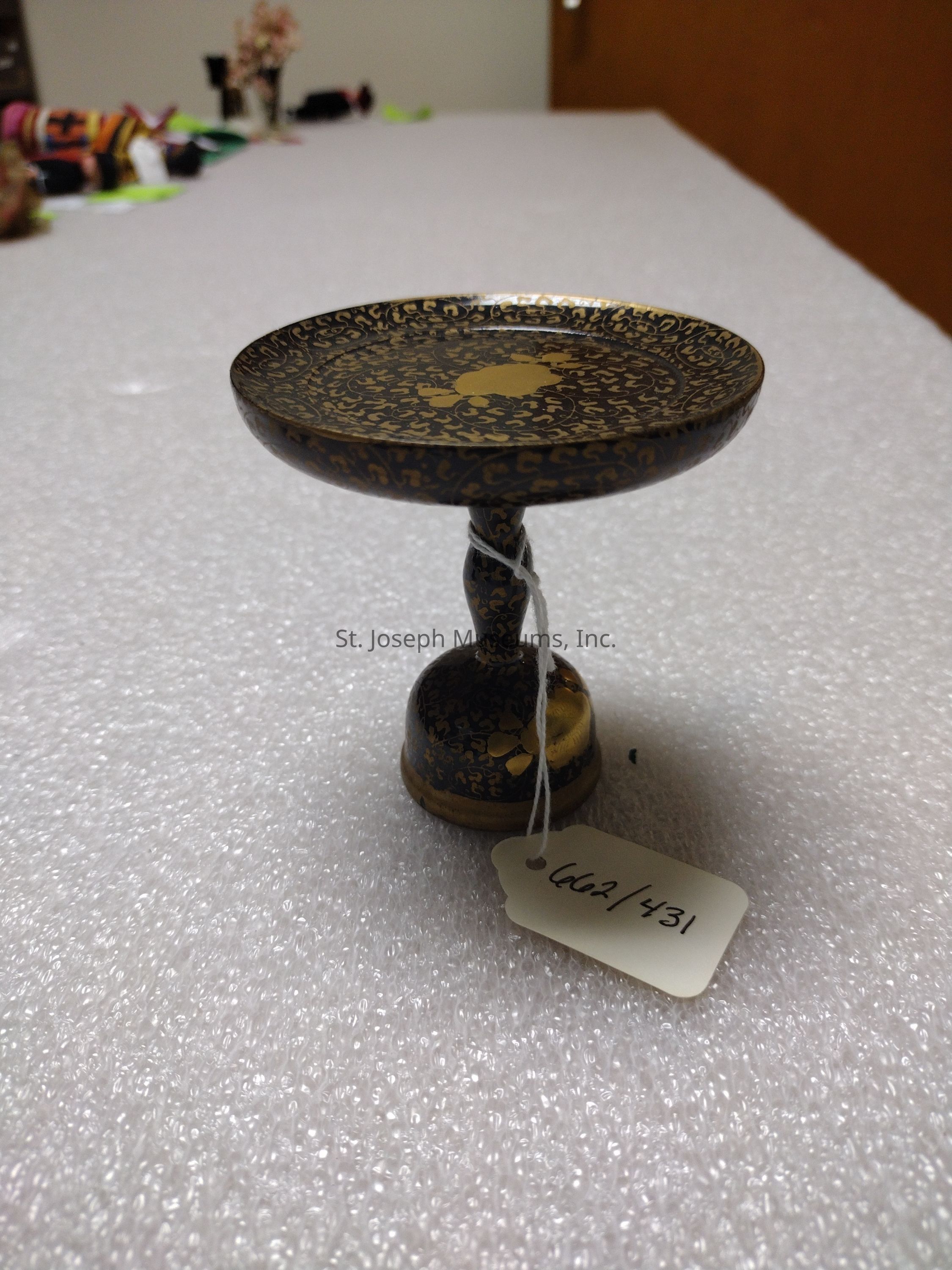 The image shows an elegant, intricately designed, decorative pedestal bowl featuring a dark base color with a detailed golden pattern covering its surface, supported by a gracefully curved stem leading to a wide, flat, round top. The pedestal rests on a textured surface that resembles bubble wrap, suggesting it might be prepared for transport or display. Attached to the bowl's stem is a small, rectangular tag with the handwritten note "662/431," possibly indicating an inventory or catalog number. In the background, there are colorful, blurred objects lined up along the edge of the table, possibly other decorative items or crafts, along with a door suggesting the setting is indoors.
