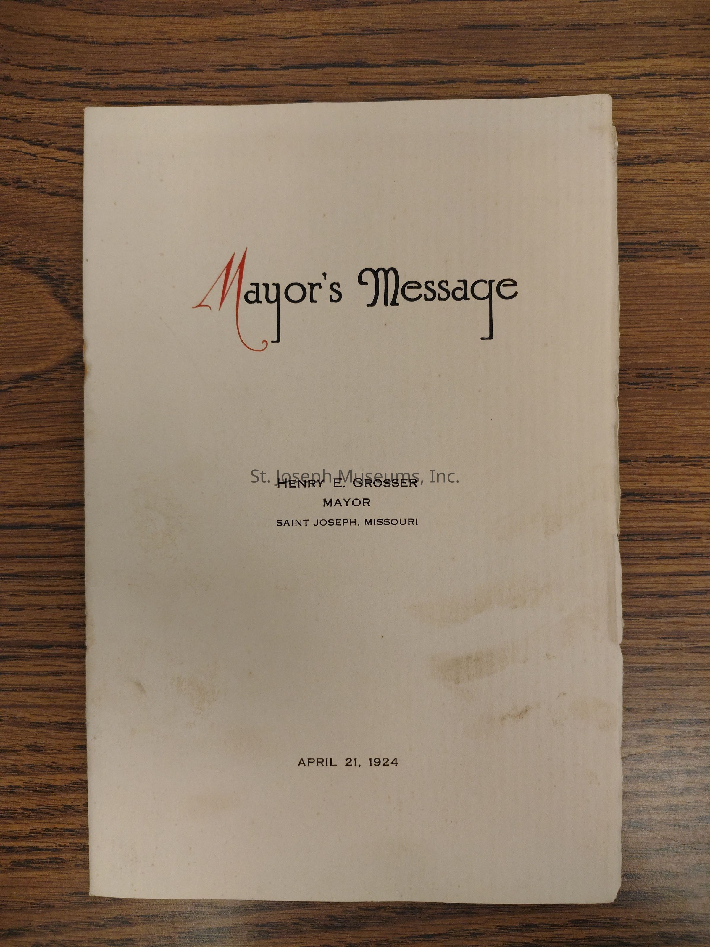 The image depicts the cover of a historical document titled "Mayor's Message," printed in a formal, elegant font with the word "Mayor's" in red and "Message" in black on an off-white, slightly aged paper. It announces the communication of Henry E. Grosser, identified as the Mayor of Saint Joseph, Missouri. The date of the message is clearly indicated at the bottom as April 21, 1924. The paper is placed on a wooden surface, showing some marks and signs of wear, suggesting its age. The text on the cover is centered and presented in a straightforward, uncluttered manner, emphasizing the significance and formality of the document. The overall appearance suggests it is an official document likely intended for public communication or record-keeping from the early 20th century, reflecting historical civic administration and tradition.