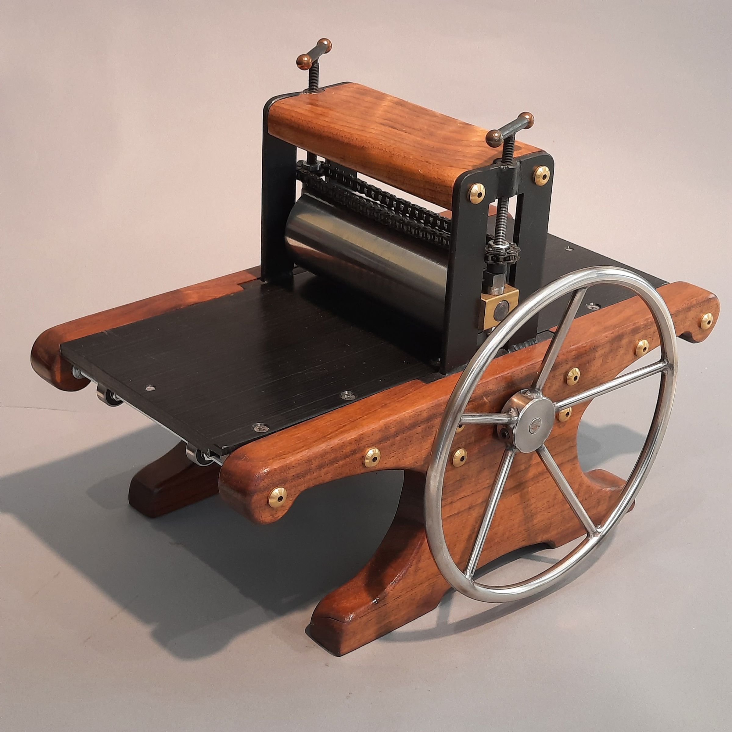 Mini Etching Press MEP6273: Most unique are the side plates are made of polished black walnut attached to the steel body with eight brass barrel cap nuts. The rack-driven bed is acetal, a plastic of hardness similar to polycarbonate measuring 3/8” thick, 6” wide and 14 1/2” long. The 8” stainless steel wheel drives the gear chain system and driving shaft with a spur gear meshes with the bed’s rack gear. The top and bottom rollers are 1 1/2” steel on roller bearings. Synchronized pressure screws raise the top roller and the synchronizing chain keeps the top roller level, assuring uniform printing pressure across the width of the plate. The press is designed to produce prints from relief plates from 1/8” to type-high blocks. Thick intaglio plates present challenges, therefore 22 to 35 gauge plates are recommended.