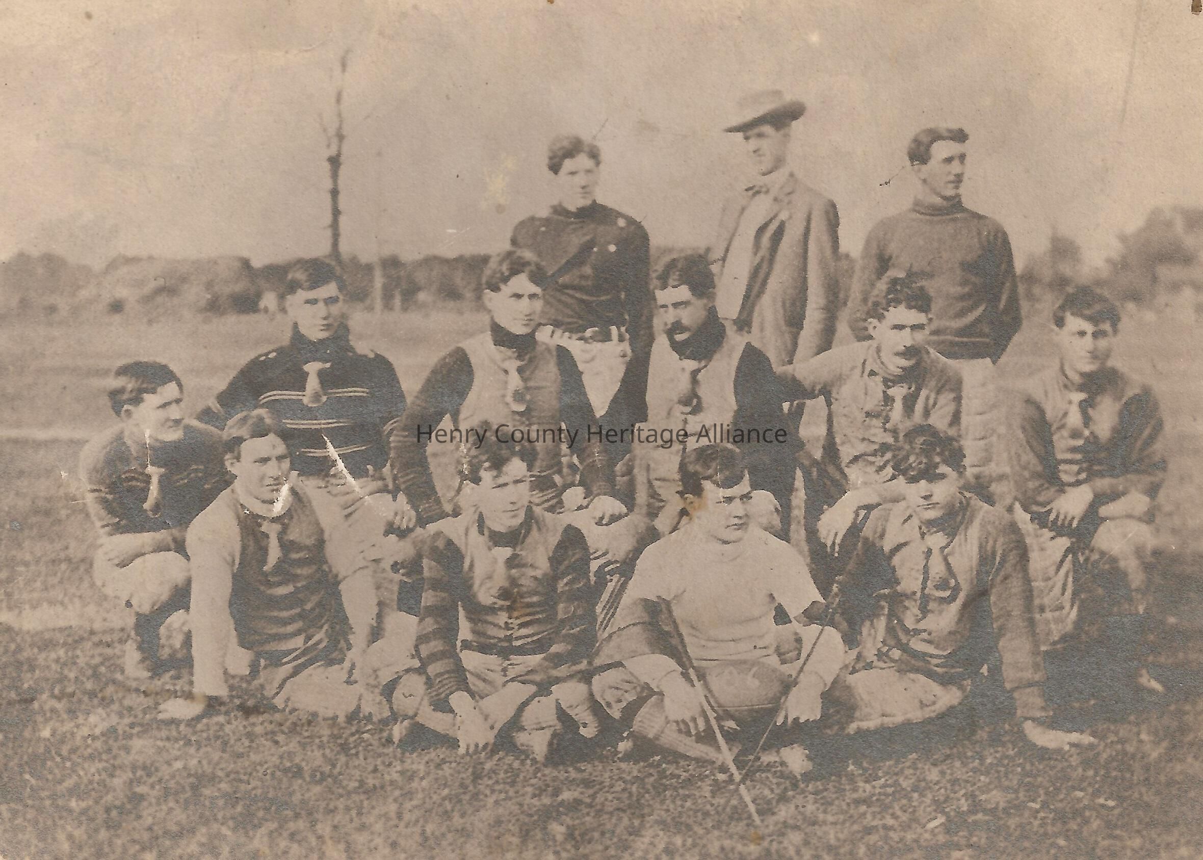 The image depicts a vintage photograph of a group of twelve men, likely a sports team from the early 20th century, posing outdoors on a grassy field. The men are arranged in three rows: the first row sitting on the ground, the second kneeling behind them, and the third standing, with one man wearing a hat and a suit, suggesting he may be the coach or manager. They are dressed in attire typical of early American football players, including padded vests and knicker-style pants. The sepia tone of the photograph adds an antique feel, emphasizing the historical context and timeless camaraderie among the team members, who display a range of expressions from serious to jovial.
