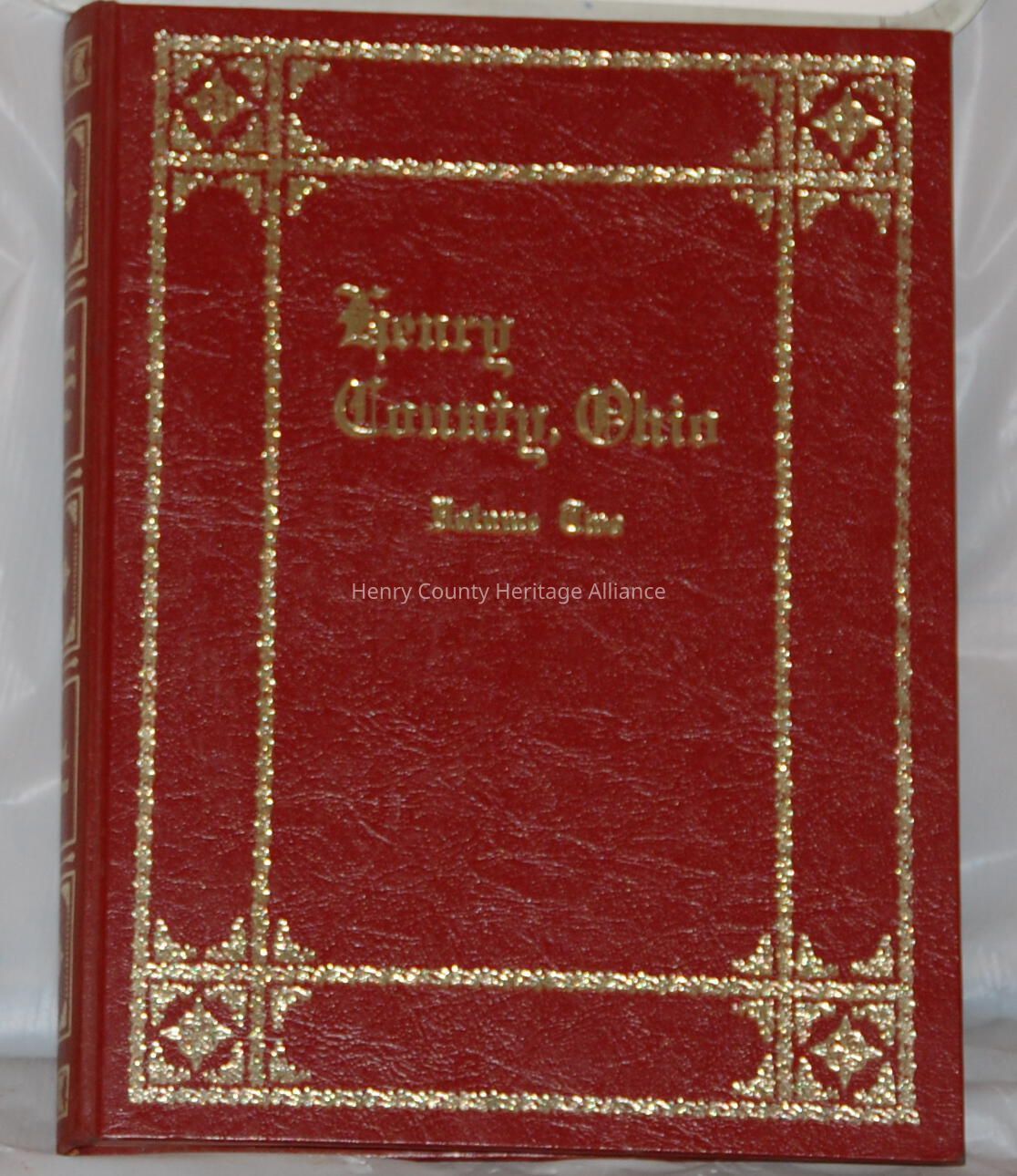 The image displays a red hardcover book with an ornate design made of gold embellishments, creating a sophisticated appearance. The title "Henry County, Ohio" is elegantly written in gold, positioned prominently in the center of the cover, with "Volume One" inscribed below it, also in gold. The book's cover features intricate decorative elements in the corners, enhancing its vintage and formal style. The spine shows similar gold detailing, complementing the overall classic and refined aesthetic of the book, suggesting it may be a historical volume or a collection of records related to Henry County in Ohio.