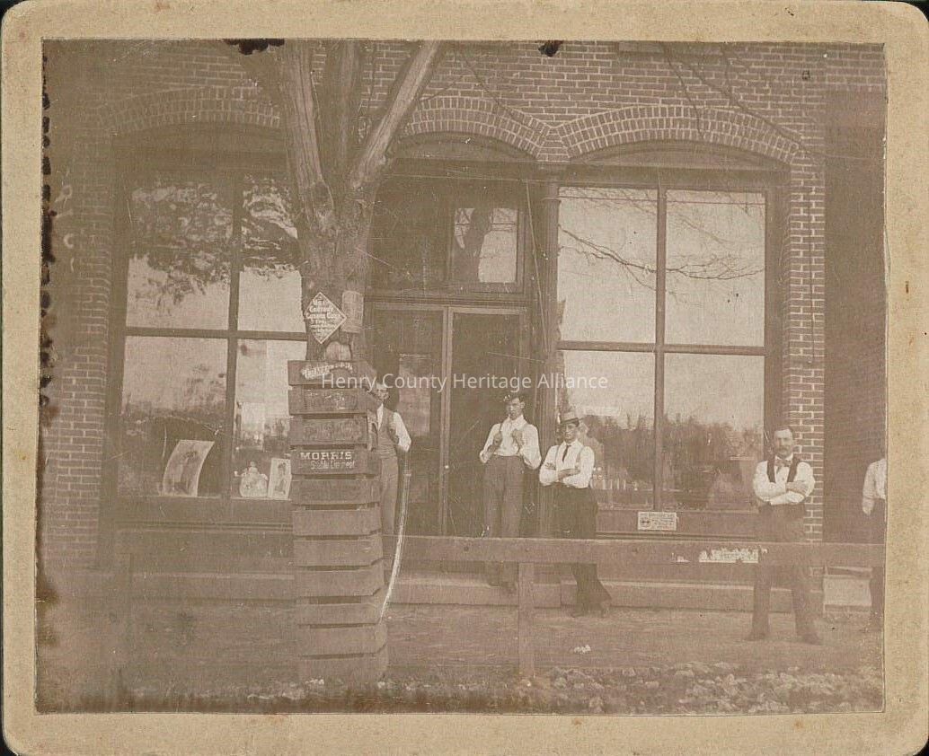 The image depicts an old sepia-toned photograph showing a group of men dressed in vintage attire standing outside Liberty Center's pharmacy with large glass windows. The men are wearing hats, dress shirts, and vests, exuding a sense of formality typical of the late 19th century. A tree stands prominently in the foreground, partially obstructing the view of the building's entrance. There is a stack of signs attached to the tree's protective barrier, with one reading "Morris" in visible letters. The windows reflect the branches of surrounding trees, adding depth to the scene. The setting suggests a small-town atmosphere, with the men appearing relaxed and engaged in conversation or perhaps posing for the photograph. The building's architectural details, including the brickwork and arched window frames, highlight the era's design preferences. Overall, the image captures a moment in time, providing a glimpse into the fashion and social dynamics of the period.