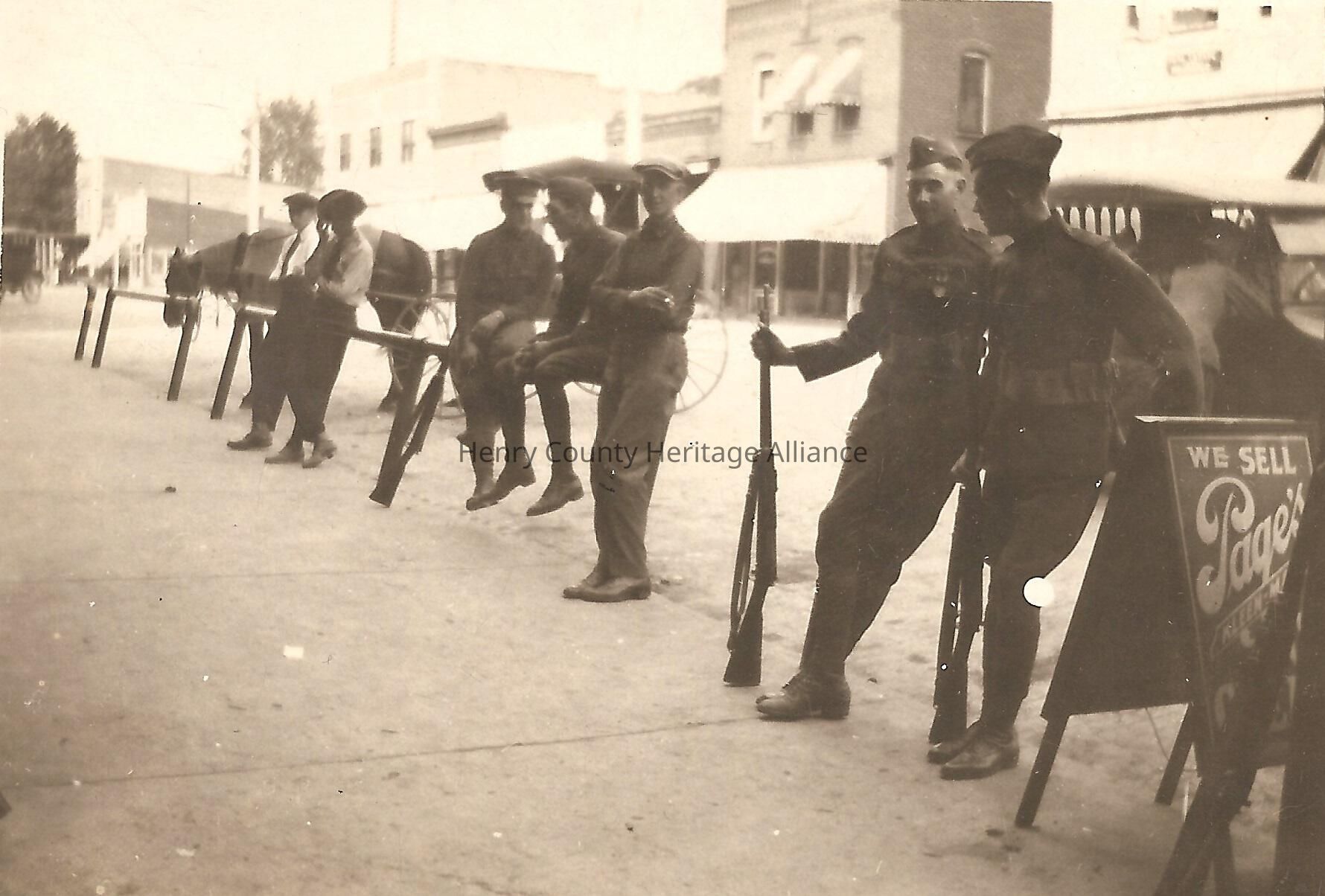 In the sepia-toned image, a group of veterans are seen casually leaning against and sitting on iron hitching posts along East Street in Liberty Center, OH, with several of them holding rifles; they are dressed in WWI clothing, including military-style uniforms and flat caps, and are positioned in front of a backdrop consisting of the east side of East Street and horse-drawn carriages, giving the scene a historical feel, while the signboard on the right reads "We Sell Page's Ice Cream." possibly referring to a store or product from that era.