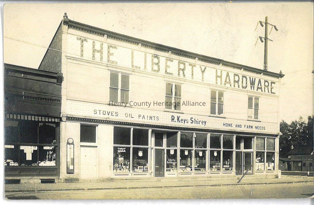 The image shows a historic, two-story building with "The Liberty Hardware" sign prominently displayed at the top, featuring large windows on the ground floor showcasing various products inside, while the sign along the building's facade advertises items like stoves, oil paints, and home and farm needs, indicating that the store is named R. Keys Shirey; in addition, the building has a classic architectural style with decorative elements along the roofline, typical of 1890s commercial establishments, and is positioned along East Street.