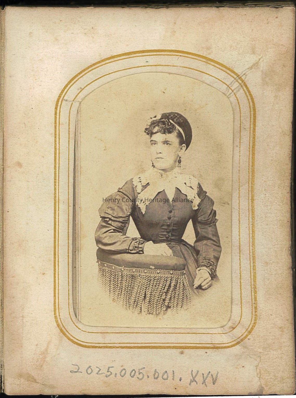 The image depicts a sepia-toned vintage photograph of a woman seated with a composed expression, dressed in a formal Victorian-era gown featuring a high collar with lace detailing, billowing sleeves, and a fitted bodice, accessorized with earrings and a decorative headpiece. The portrait is framed within an ornate border, suggesting its preservation within a period-style album or collection, evoking a sense of historical elegance and offering a glimpse into the fashion and photographic practices of the past.
