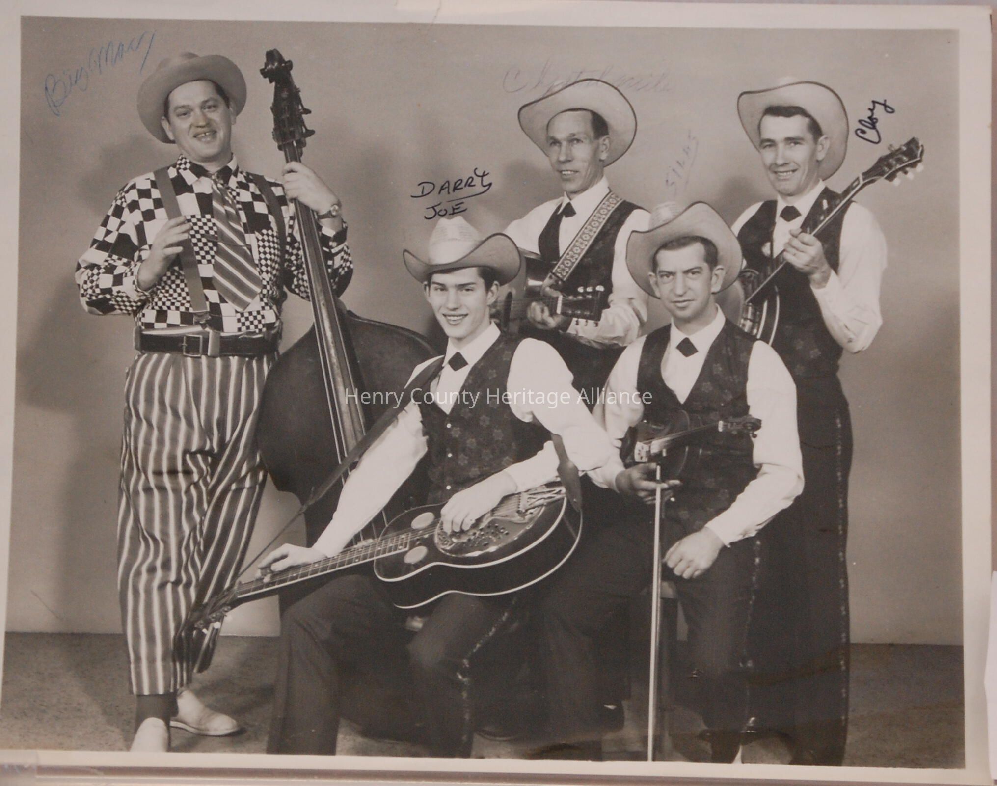 In the black-and-white image, a group of five musicians is arranged in a classic pose. They all wear matching cowboy hats and vests, complete with ties and white shirts, embodying a vintage Western-inspired style. The man on the far left stands with a large double bass, sporting a unique outfit with a checkered shirt and striped pants, adding a touch of individuality. Seated in the middle, one musician holds a guitar, while another, standing behind him, also holds a guitar. To the right, the fourth member is playing a banjo, and the final musician, seated in front, holds a fiddle. The group exudes a sense of camaraderie and musical unity, captured in an era that speaks to the roots of country or bluegrass music. Each member appears poised and focused, ready to perform, and the photograph is signed, suggesting it might have been a promotional or commemorative image for the band.
