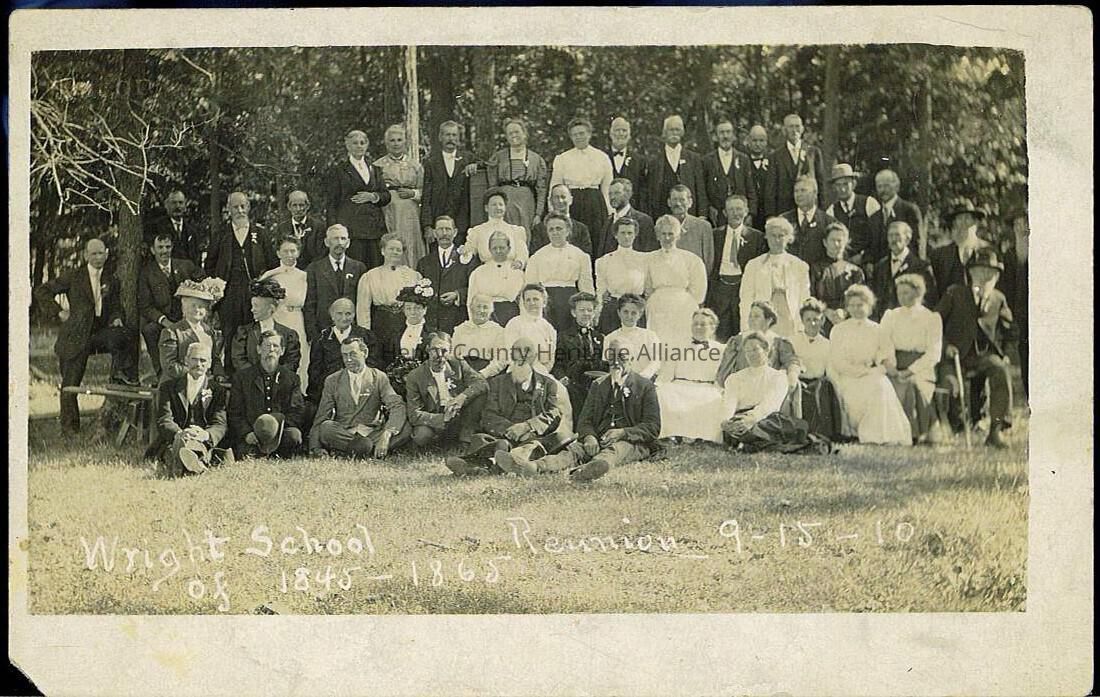 The image shows a large group of individuals gathered together outdoors, posing for a photograph that appears to be a reunion event. There are several rows of people dressed in formal attire, such as suits and long dresses, suggesting a historical or vintage setting. The backdrop includes trees, indicating that the photo was taken in a natural setting, likely a park or a garden. Written on the photograph is text identifying it as the "Wright School Reunion" and including the dates "1848-1865" as well as "9-15-16," which could represent its historical context and the reunion date. The group appears composed of a mix of men and women, with some individuals seated on the grass or benches, while others stand behind them. The expressions on their faces vary, as they gaze toward the camera, capturing a moment of shared history and possibly commemorating their connection to the school mentioned. The photo, aged and bordered, enhances its antique quality.